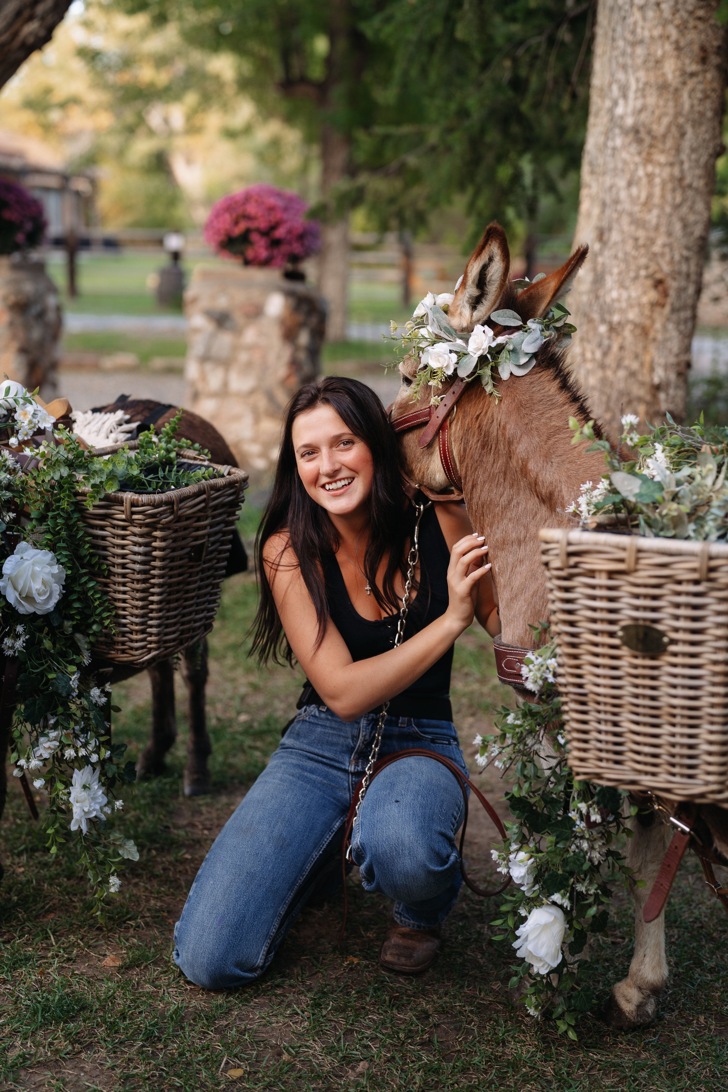 Woman kneeling next to a decorated donkey with a floral crown, smiling outdoors in a park-like setting with baskets of flowers and greenery.