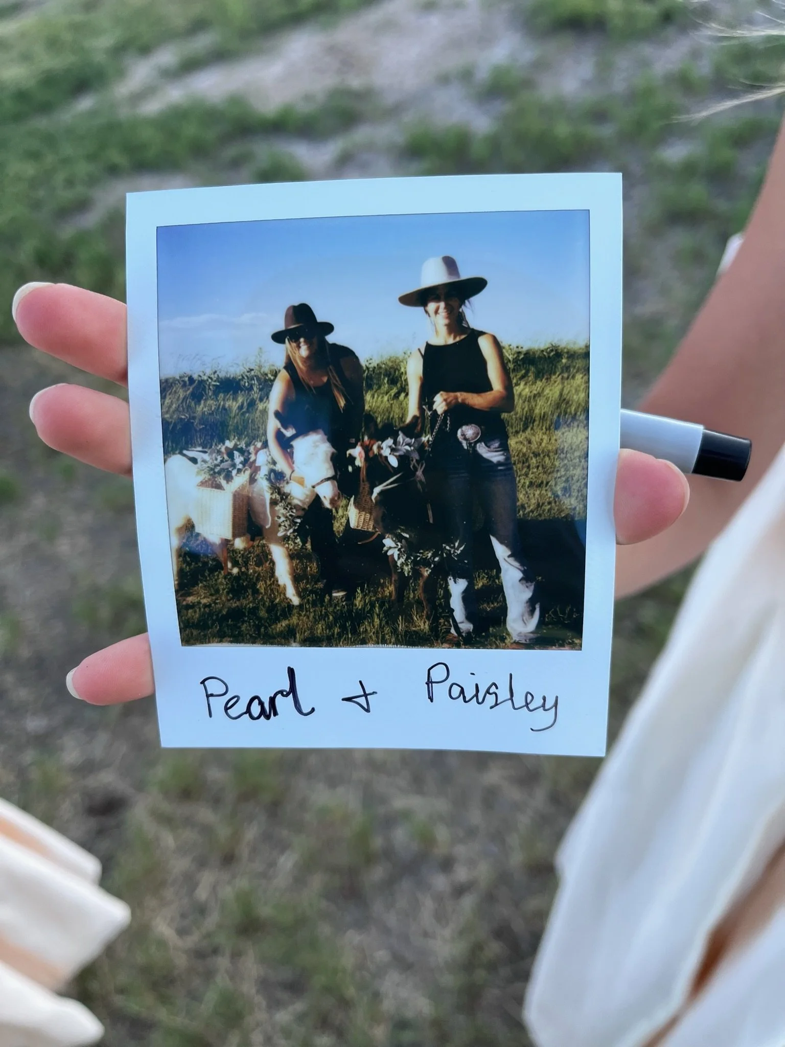 A color photograph of two women wearing black sleeveless tops and wide-brimmed hats, standing outdoors in a grassy field on a sunny day. They are smiling and holding small animals, possibly sheep or goats, with a handwritten note at the bottom of the photo that reads "Pearl + Paisley." The background shows a clear blue sky and some plants or shrubs.