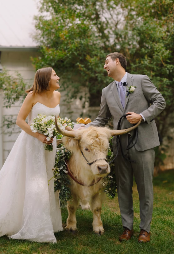A bride and groom kiss during a wedding ceremony outdoors, with a cow decorated with flowers between them.