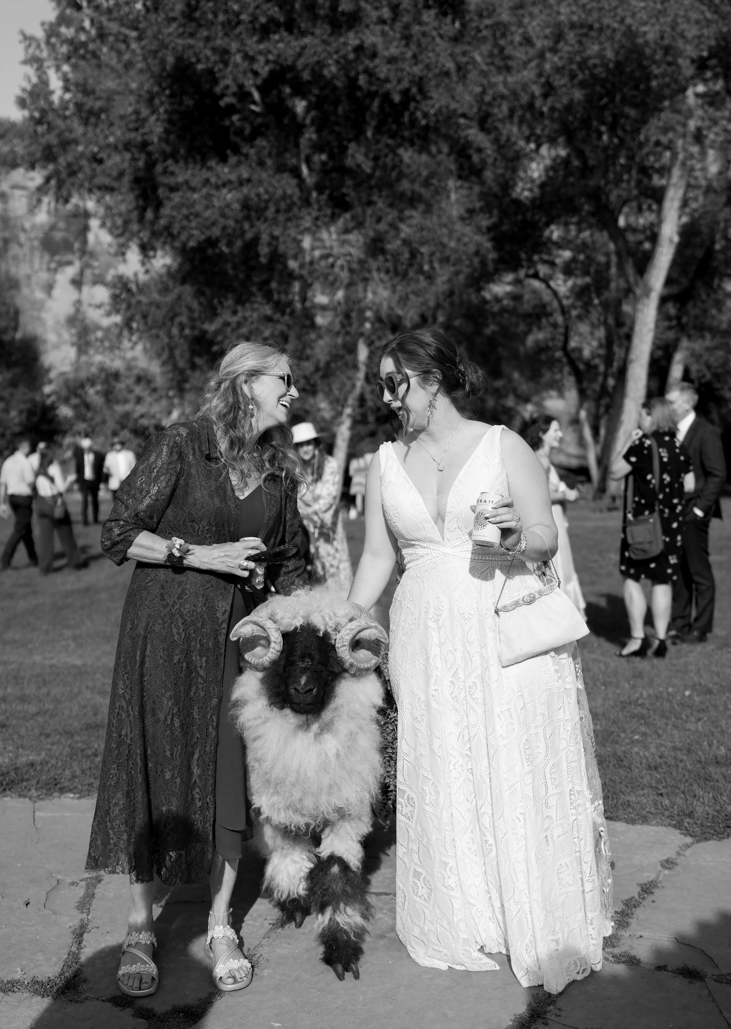 Two women in conversation next to a sheep with curled horns at an outdoor event, others in the background, some holding drinks, under trees on a sunny day.