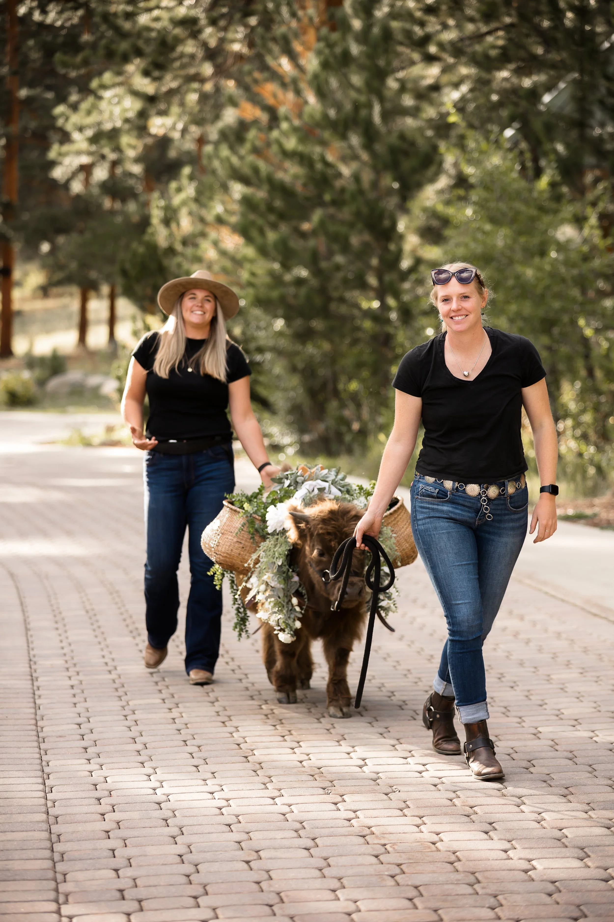 Two women, one holding a small brown cow calf in a basket decorated with flowers, walking on a brick path outdoors surrounded by trees.