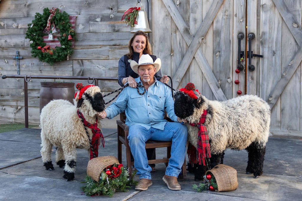 A man in a cowboy hat and blue shirt sitting on a wooden chair, with two sheep wearing red scarves and Christmas hats beside him. A woman in a denim jacket stands behind, smiling, holding the man's shoulder. The background features a wooden barn door with festive decorations including a wreath, bells, and Christmas ornaments.
