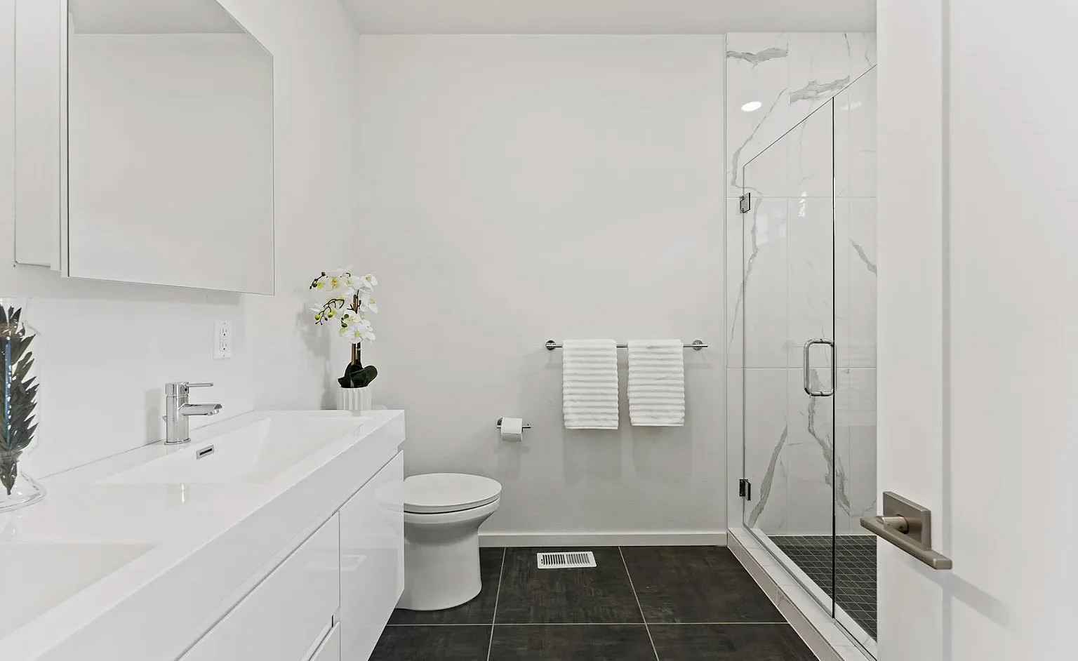 Modern bathroom with white vanity, large mirror, toilet, glass-enclosed shower, and tiled floor.