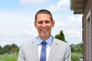 Dr Robert Pieters in a light gray suit and blue tie standing outdoors near a brick building with trees and blue sky in the background.