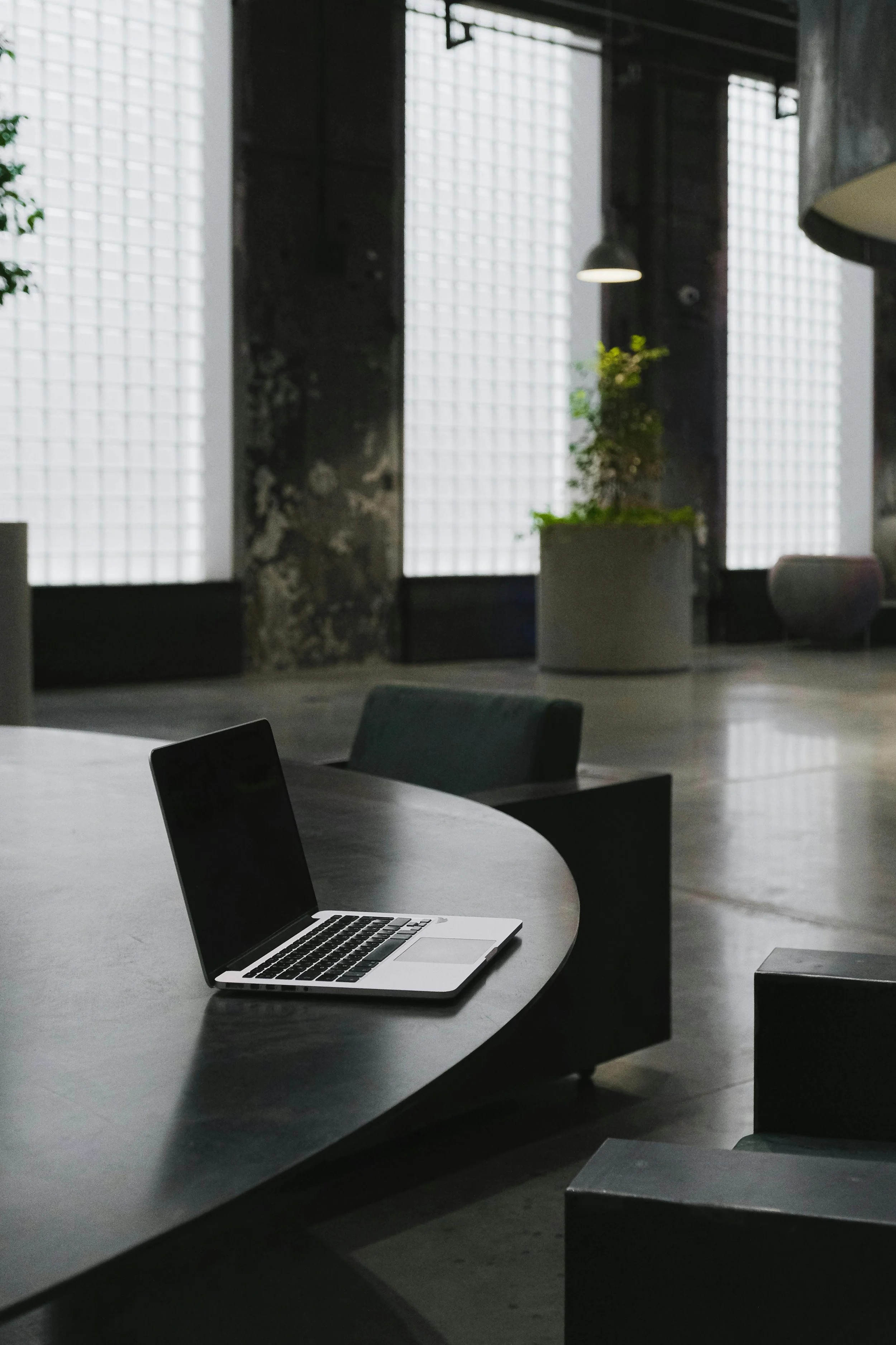 Open laptop on a round table in a modern industrial-style room with large windows and plants.
