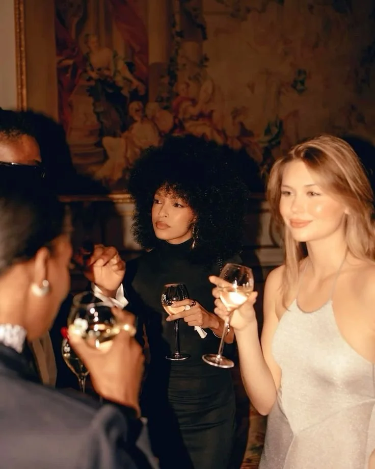 People socializing at a formal event, holding wine glasses, with elegant clothing and a decorative backdrop.