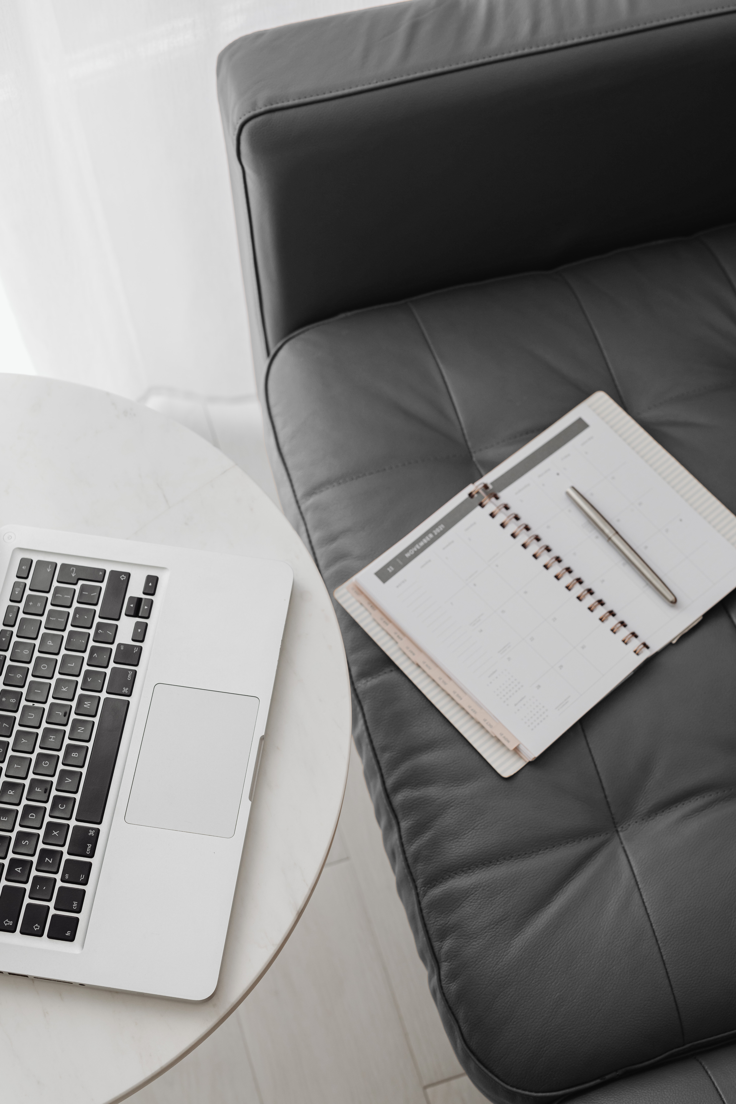 Open laptop on a marble table next to a leather couch with a planner and pen.