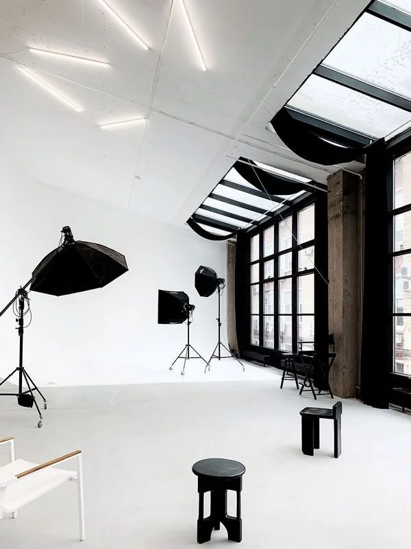 Photography studio with large windows, lighting equipment, and black stools with a white chair.