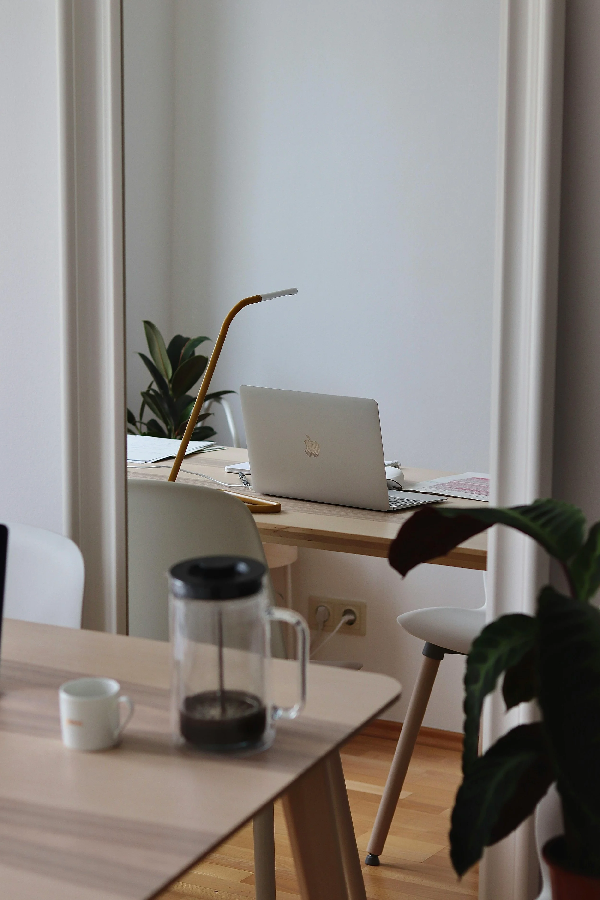 Minimalist workspace with a desk lamp, laptop, and coffee press on a wooden table, surrounded by plants.