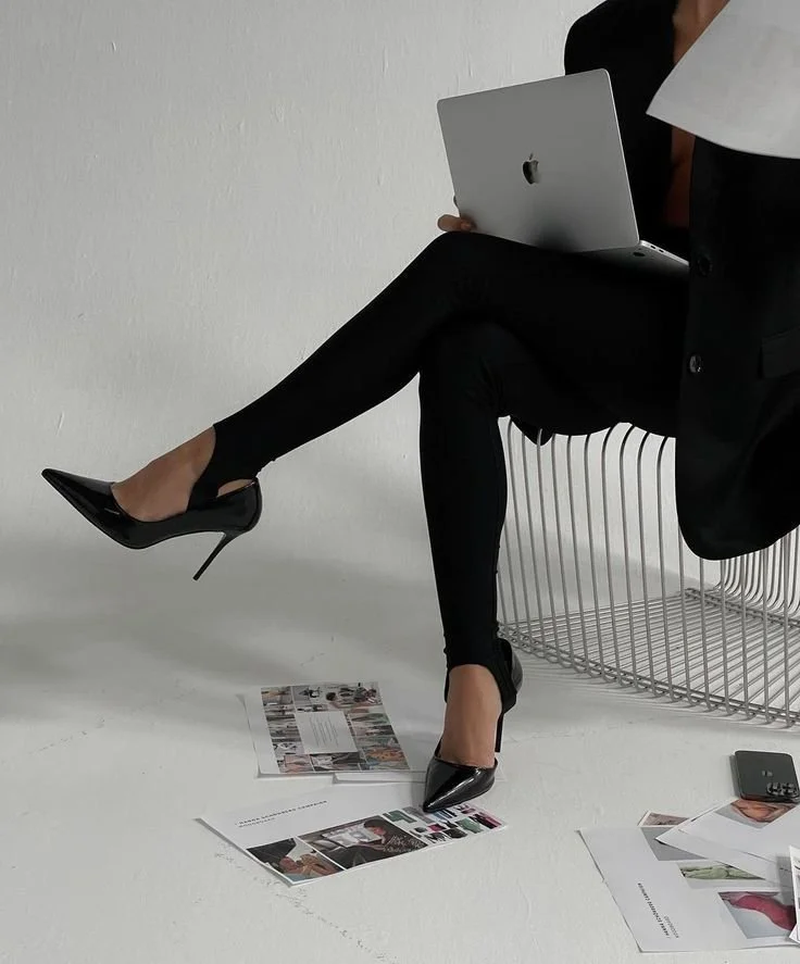 Person wearing black outfit with heels holding a laptop on a wire chair, surrounded by scattered documents on the floor.