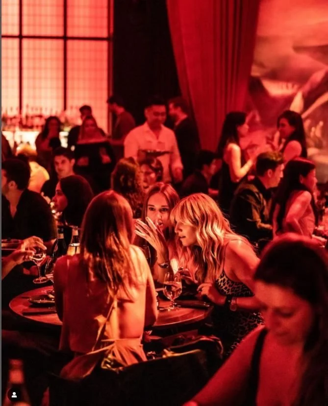 People socializing in a dimly lit bar with red lighting, sitting at tables, enjoying drinks and conversations.