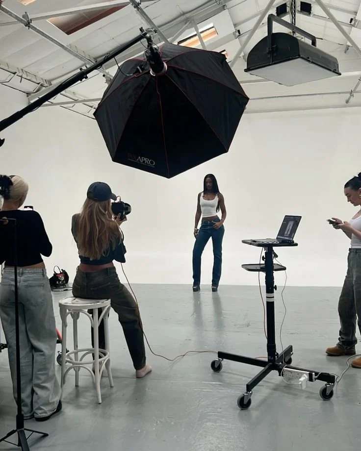 Photography studio setup with a model, photographer, and crew. Large softbox lighting overhead, laptop on stand, and photographer capturing the model. White backdrop and high ceilings overhead.