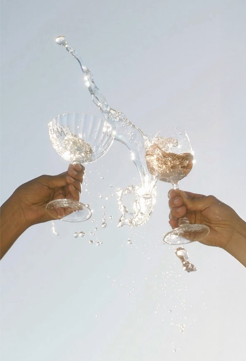 Two people clinking wine glasses with liquid splashing in the air, set against a clear blue sky.