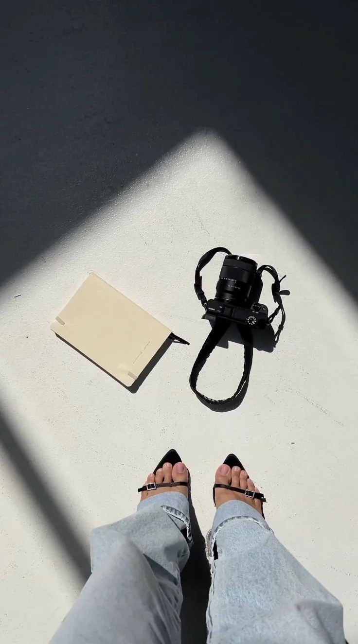 Person wearing denim jeans and black sandals standing on light-colored floor with a camera and a notebook nearby in sunlight.