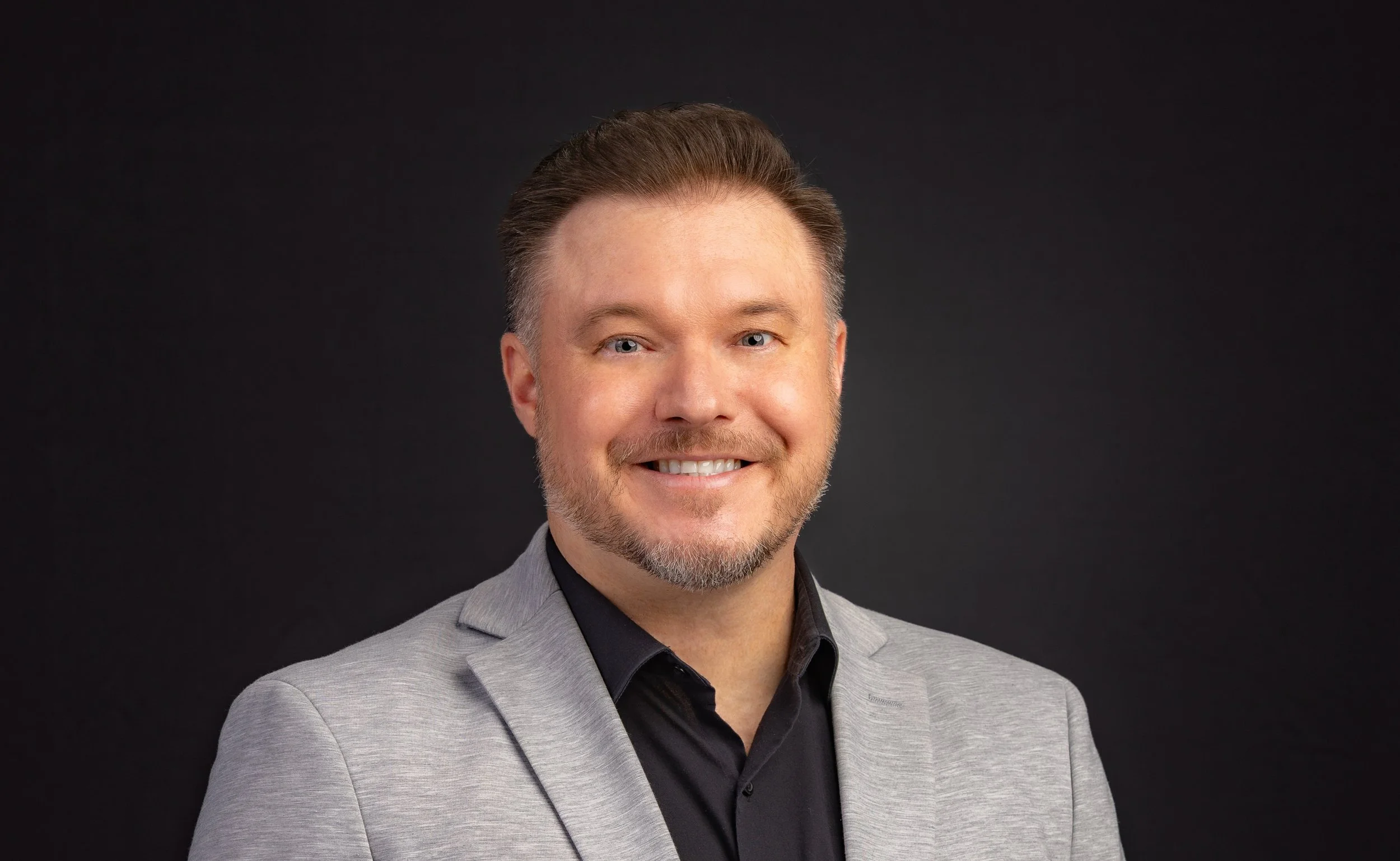 Headshot of a man with short, styled brown hair, light beard, and blue eyes, wearing a light gray suit jacket and black collared shirt, smiling against a black background.