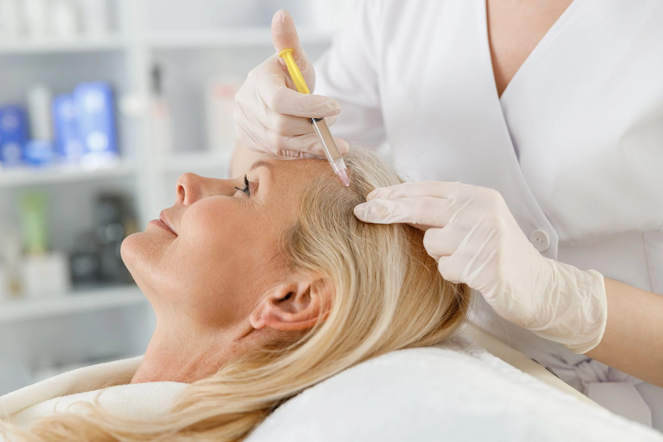 Woman lying on a treatment bed receiving a facial treatment with a handheld device from a medical professional wearing blue gloves, with medical equipment in the background.
