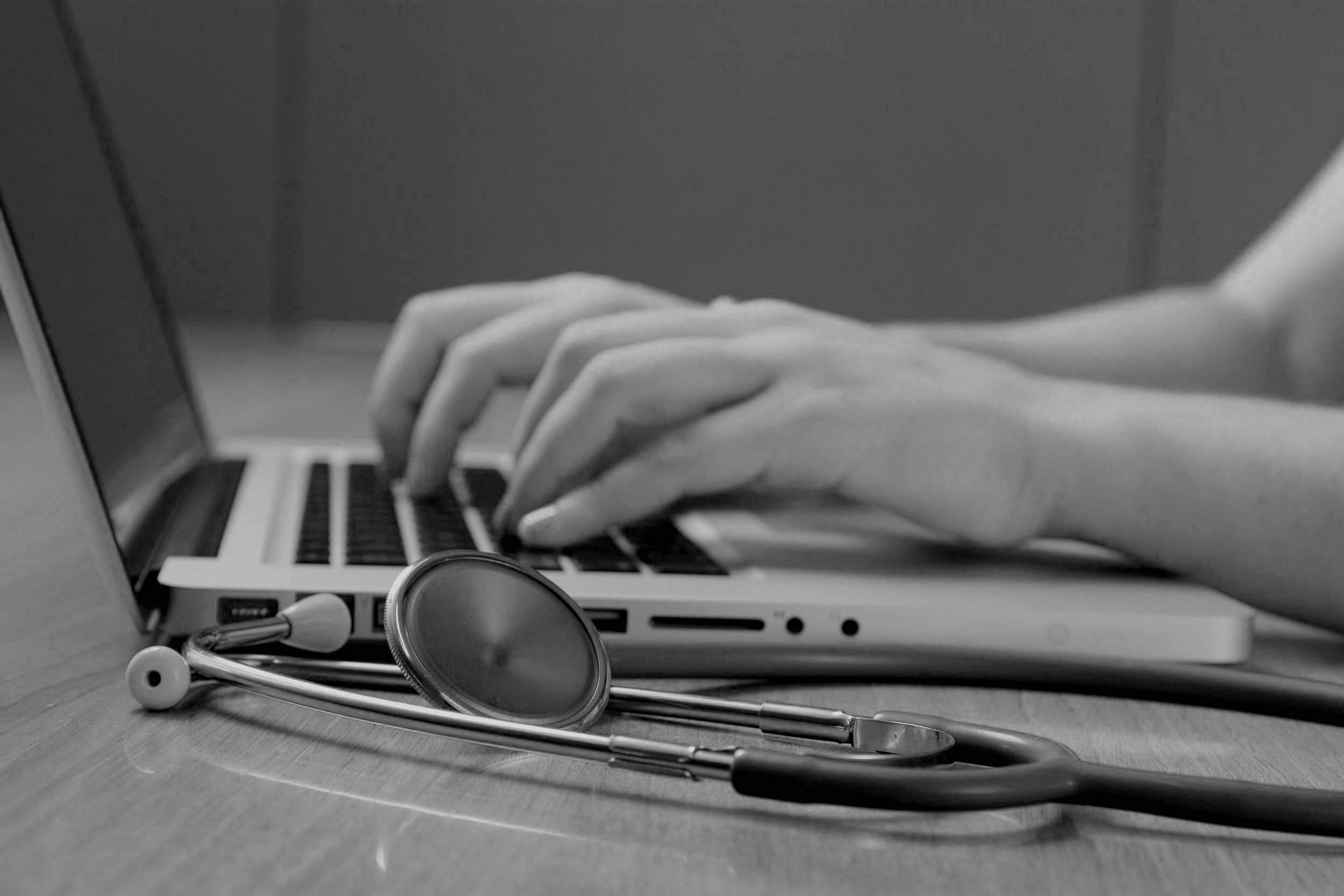 Close-up of a person's hands typing on a laptop keyboard with a stethoscope resting on the desk in front of the laptop.