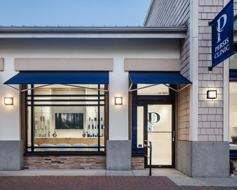 Exterior view of a clinic named Persis Clinic with blue awnings, large windows, and a glass door, located in a shopping plaza with brick and siding walls.