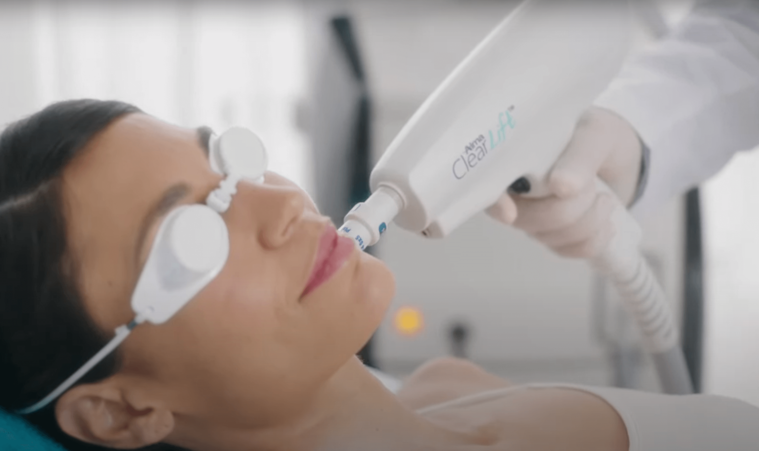 A woman receiving a cosmetic laser treatment on her lips with a device labeled 'Clear + Brilliant' while wearing protective eye shields in a clinical setting.