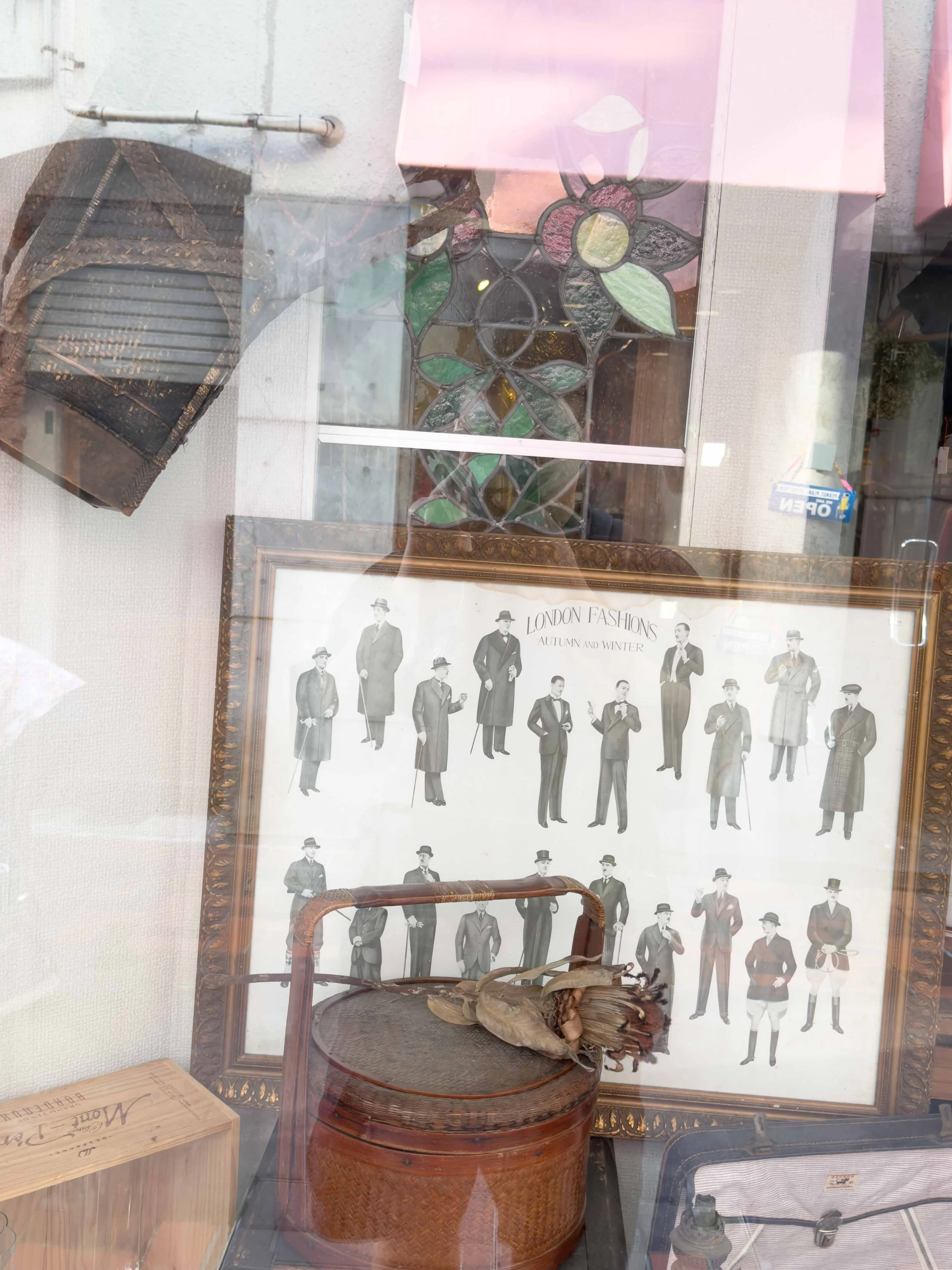 ショーウィンドウに飾られたクラシックな洋服のイラストの額縁と古いラッパの置物、ステンドグラスの花と木製の箱が見える