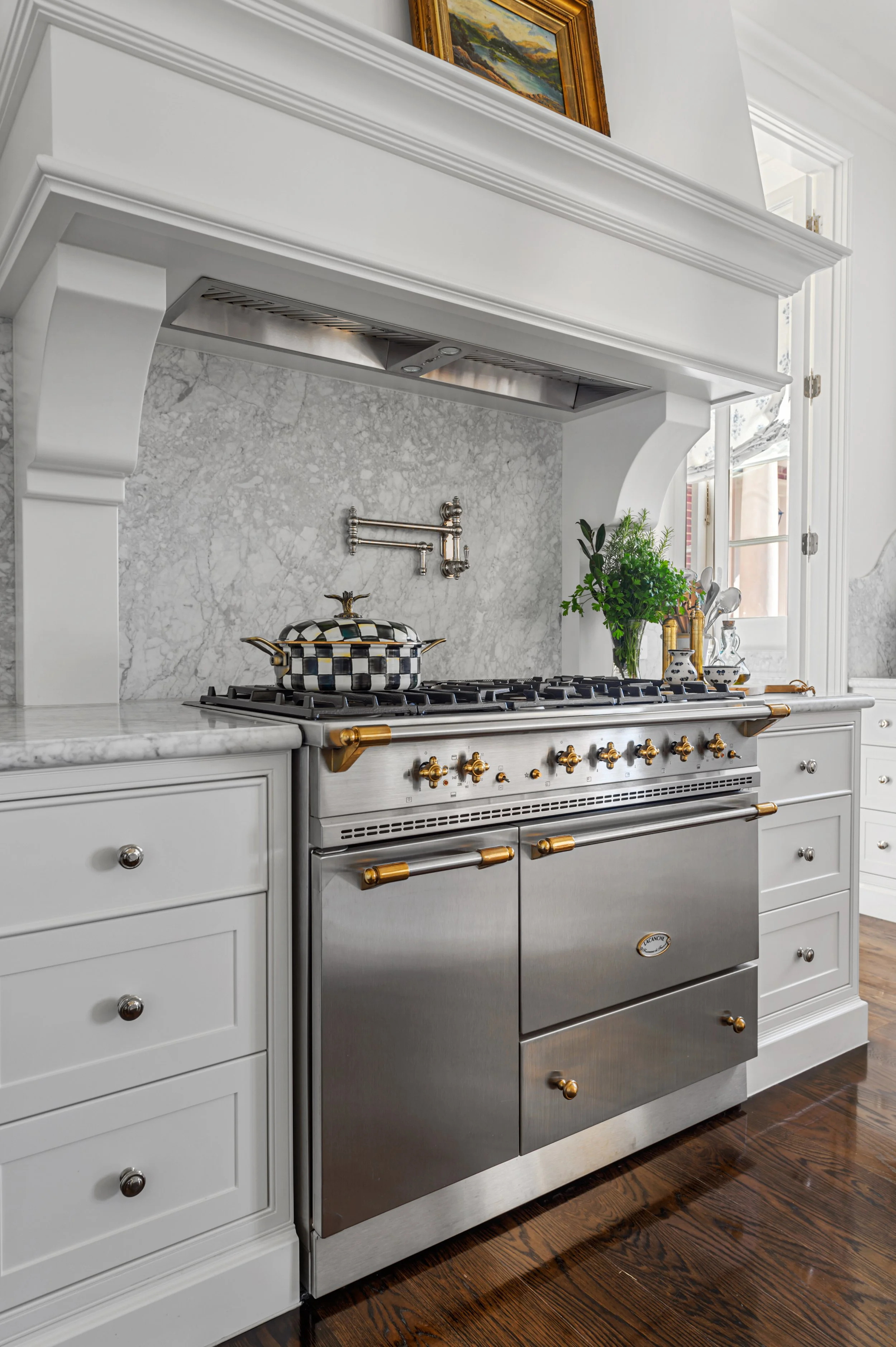 A white kitchen with a built-in stove and oven, featuring brass handles, a marble backsplash, and a window with natural light. Decorative items and a plant are on the counter.