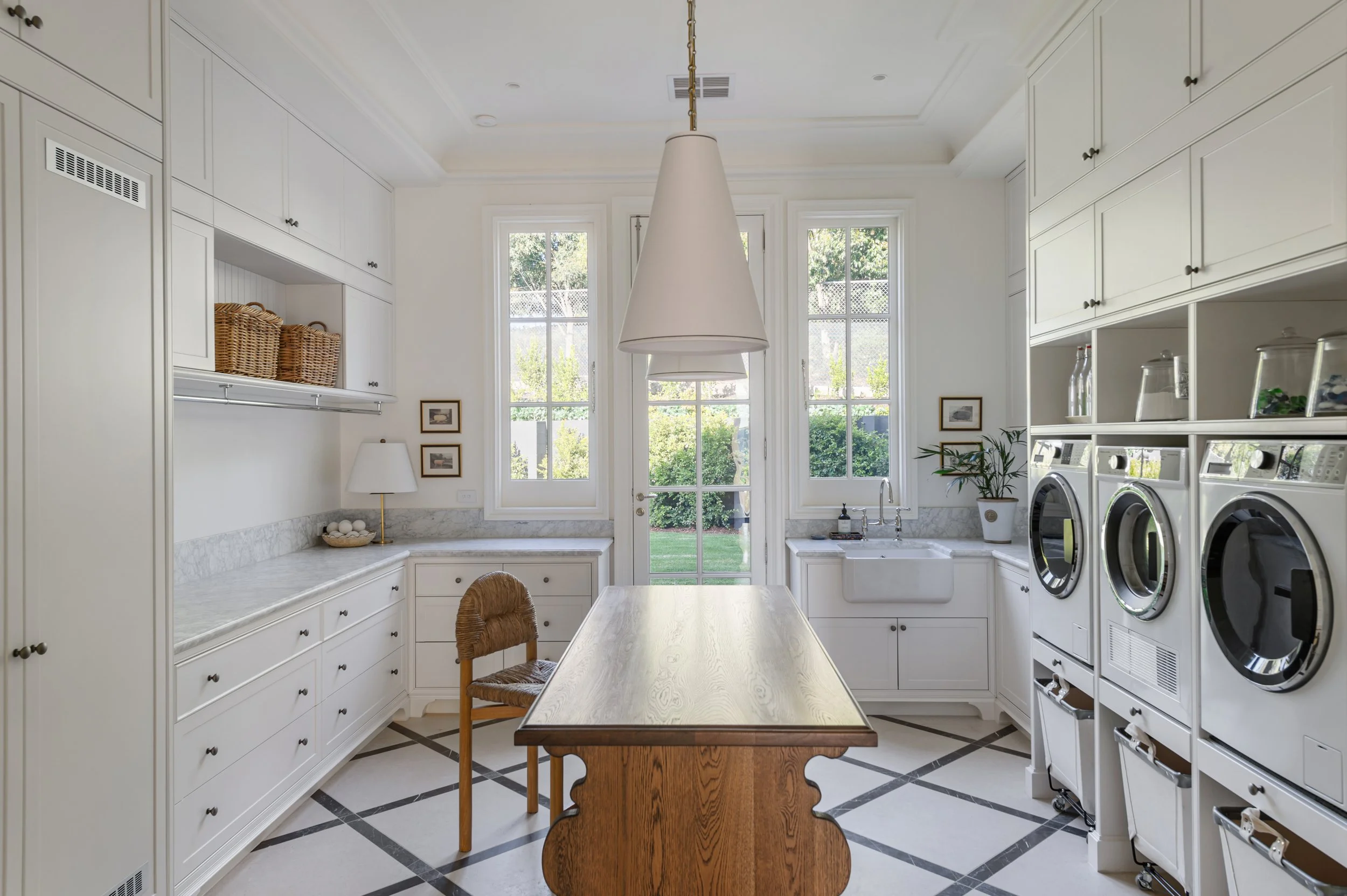 Laundry room with white cabinets, washer and dryer, a wooden table, and large windows framed by white trim.