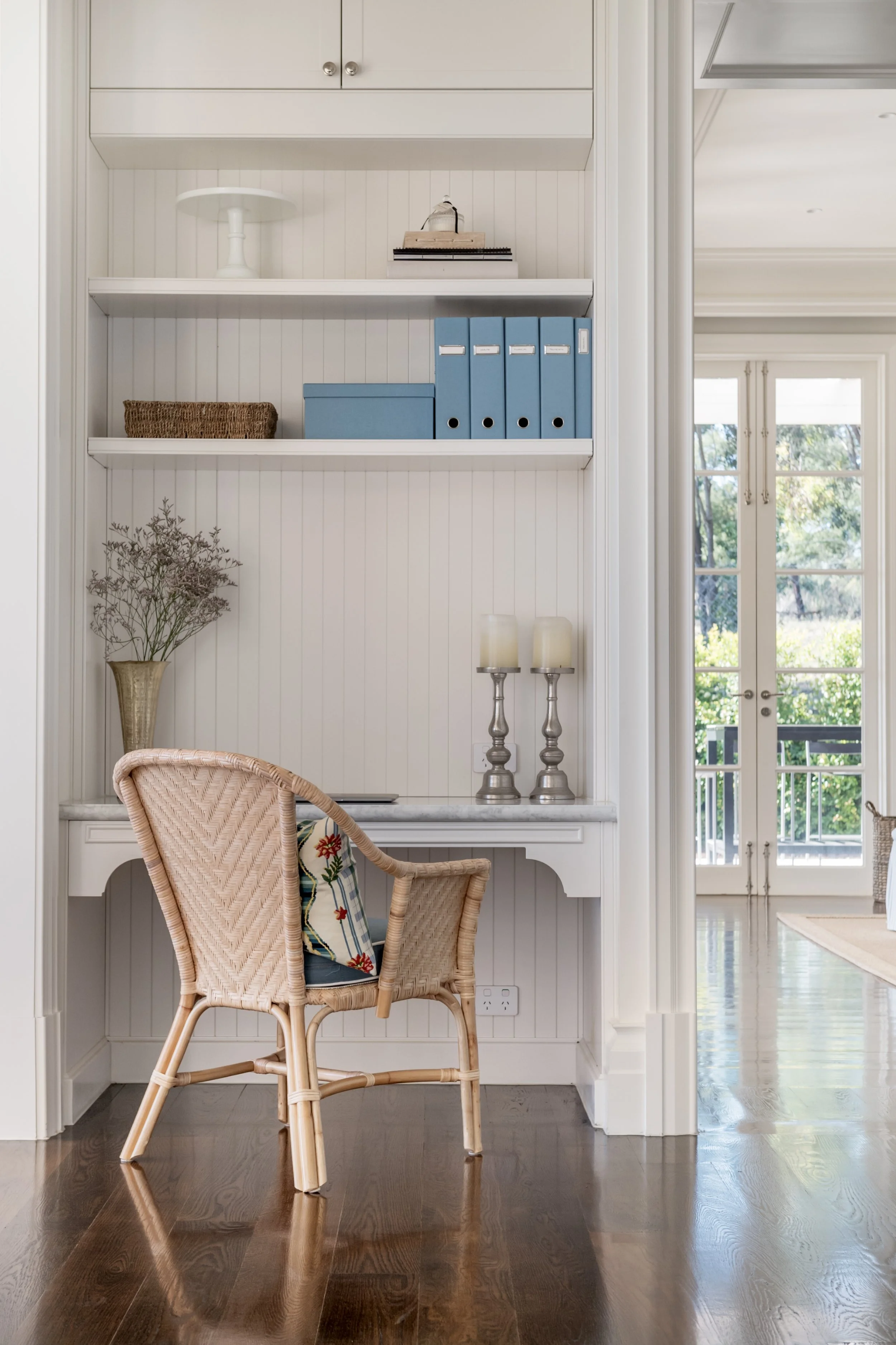 A built-in white desk with shelves above, a wicker chair with a cushion, a vase with dried flowers, and candles, with large outdoor windows in the background.
