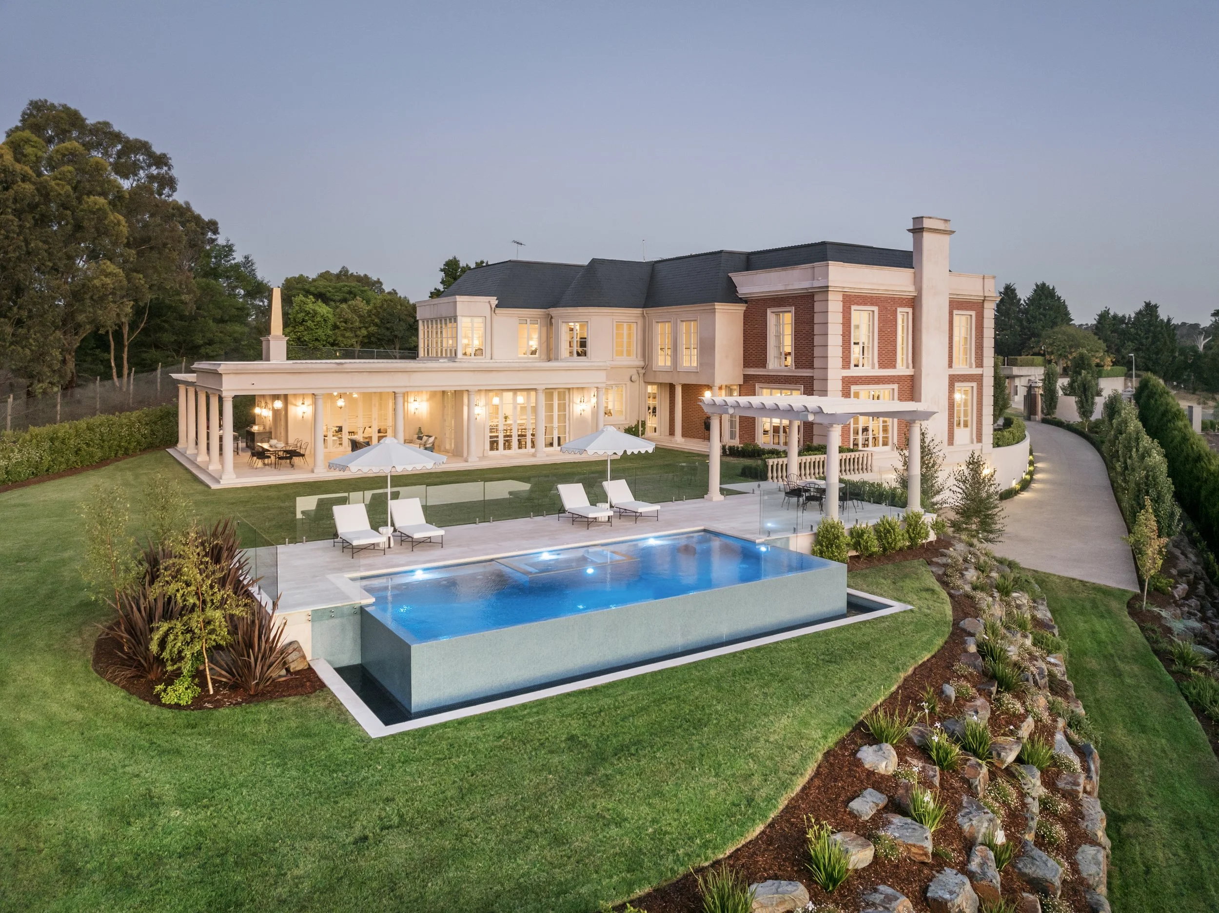 Large luxurious house with illuminated interior, swimming pool with underwater lighting, outdoor loungers, umbrellas, patio area, mature landscaping, curved driveway, and scenic hillside yard.