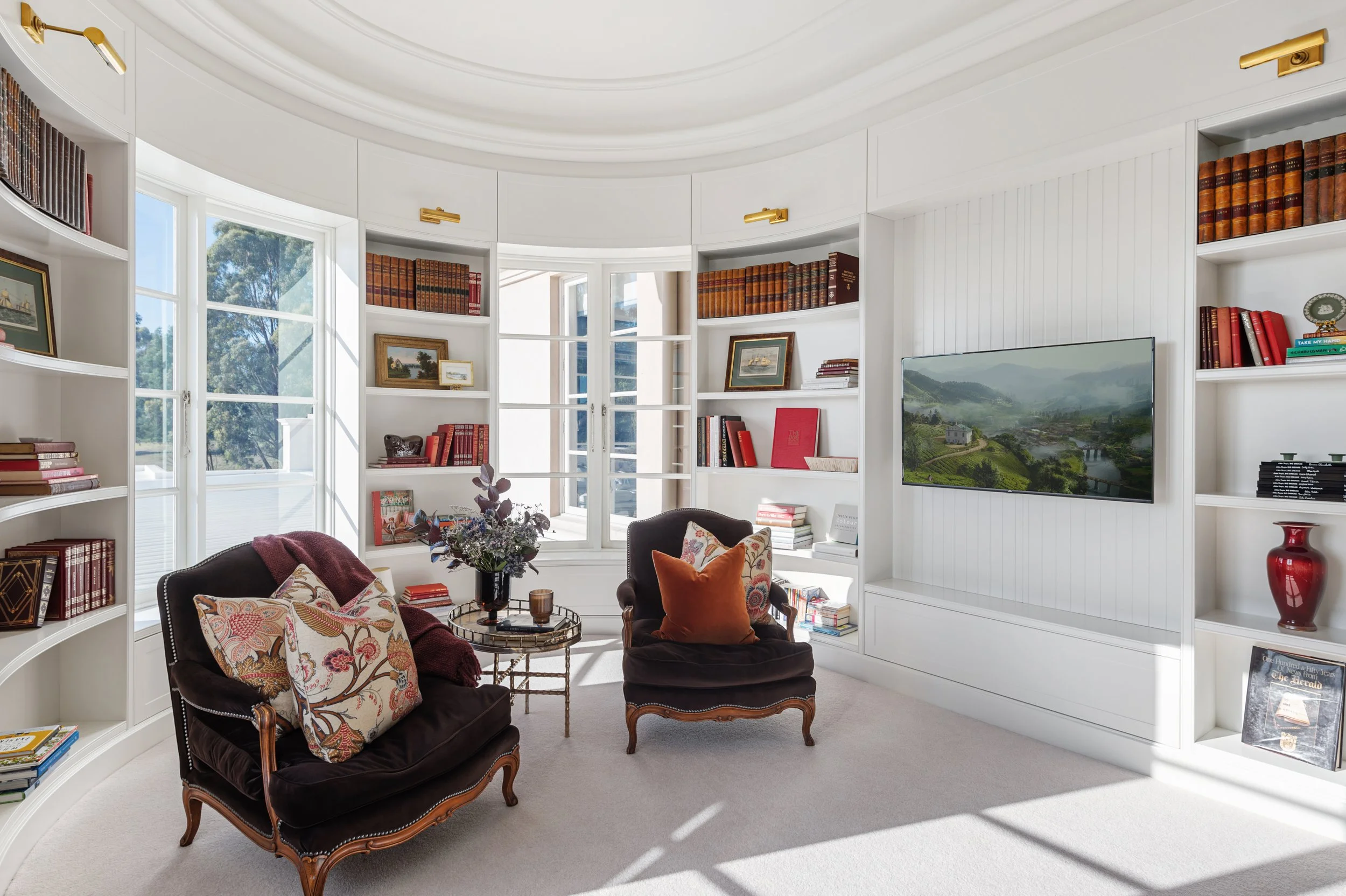 A bright, cozy living room with white curved bookshelves filled with books and decorative items, a large window with natural light, two upholstered armchairs with floral and orange cushions, a small round glass table with a vase of flowers, a wall-mo
