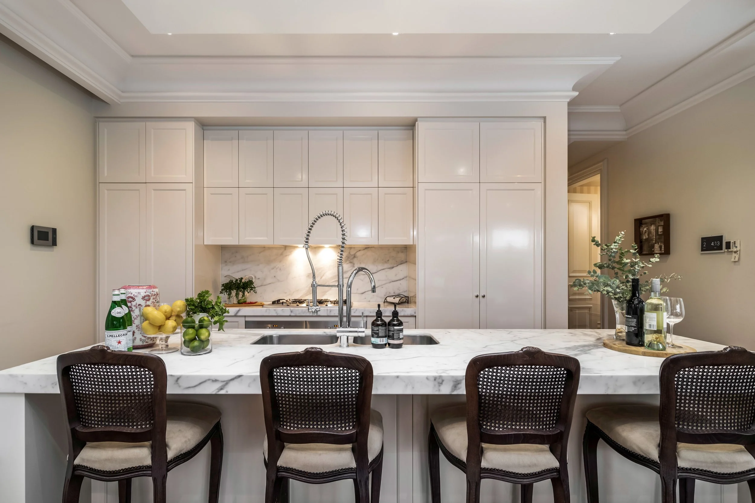 Modern kitchen with white cabinetry, marble countertops, and a sink with a high-arc faucet. Bar stools with dark wood frames and beige cushions are at the counter. Decor includes bottles of wine and spirits, a glass of wine, a fruit bowl with lemons 