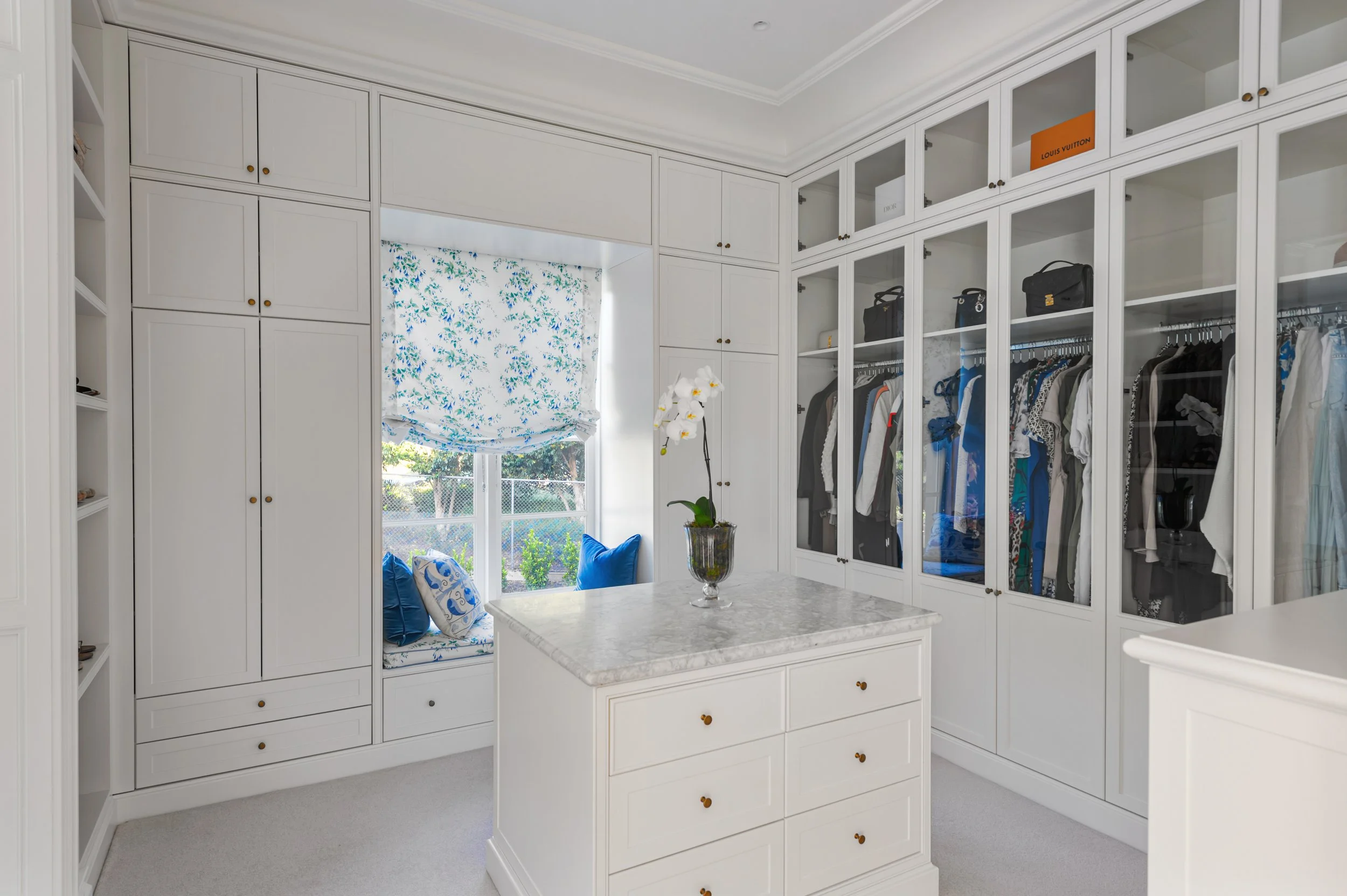 White walk-in closet with built-in cabinets, glass-front wardrobes, and a marble-topped central island. There is a window with a blue floral Roman shade and a window seat with blue and patterned pillows. A potted orchid is on the island.