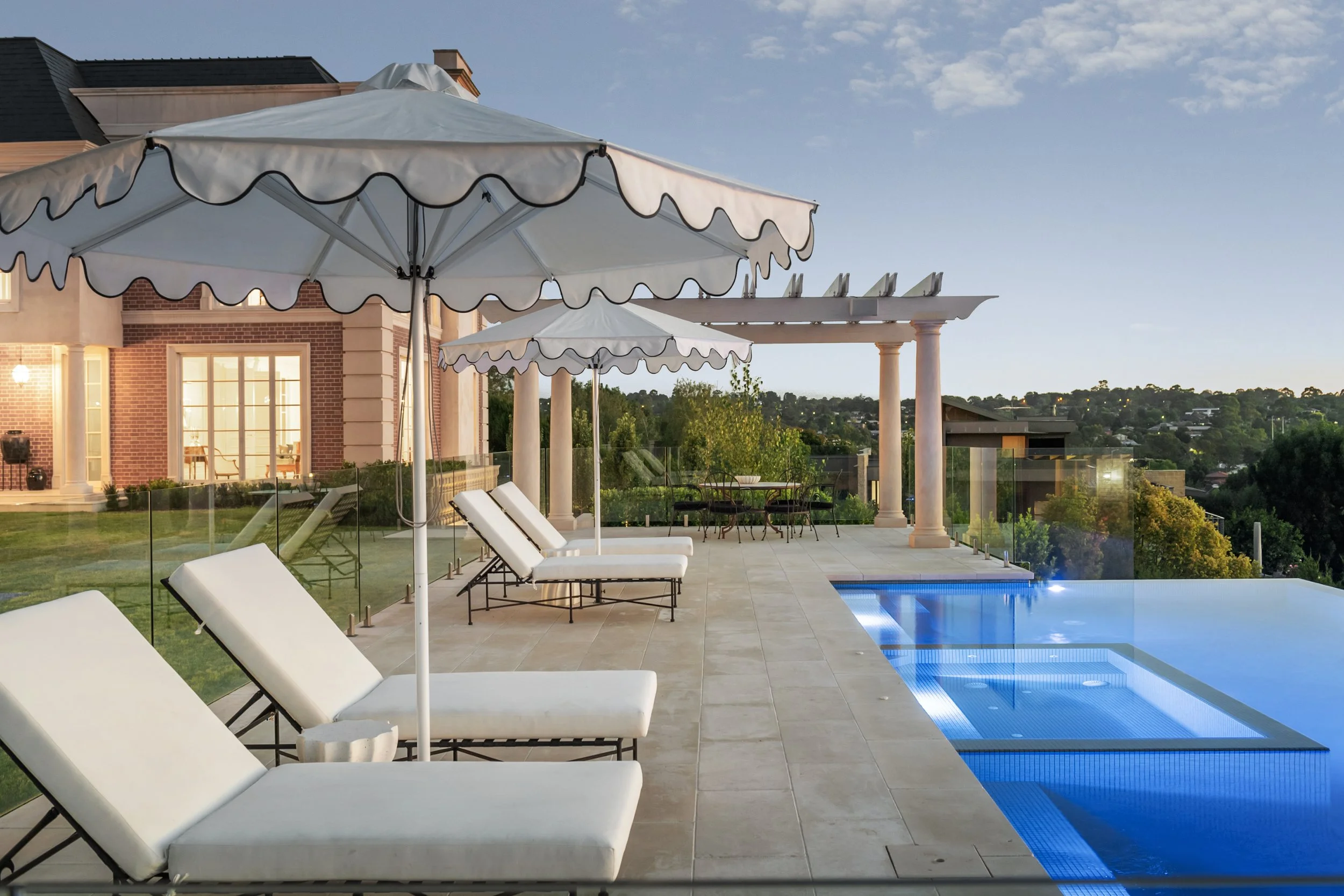 A luxurious outdoor pool area at sunset with four lounge chairs and umbrellas, a house in the background, and scenic hills and trees.