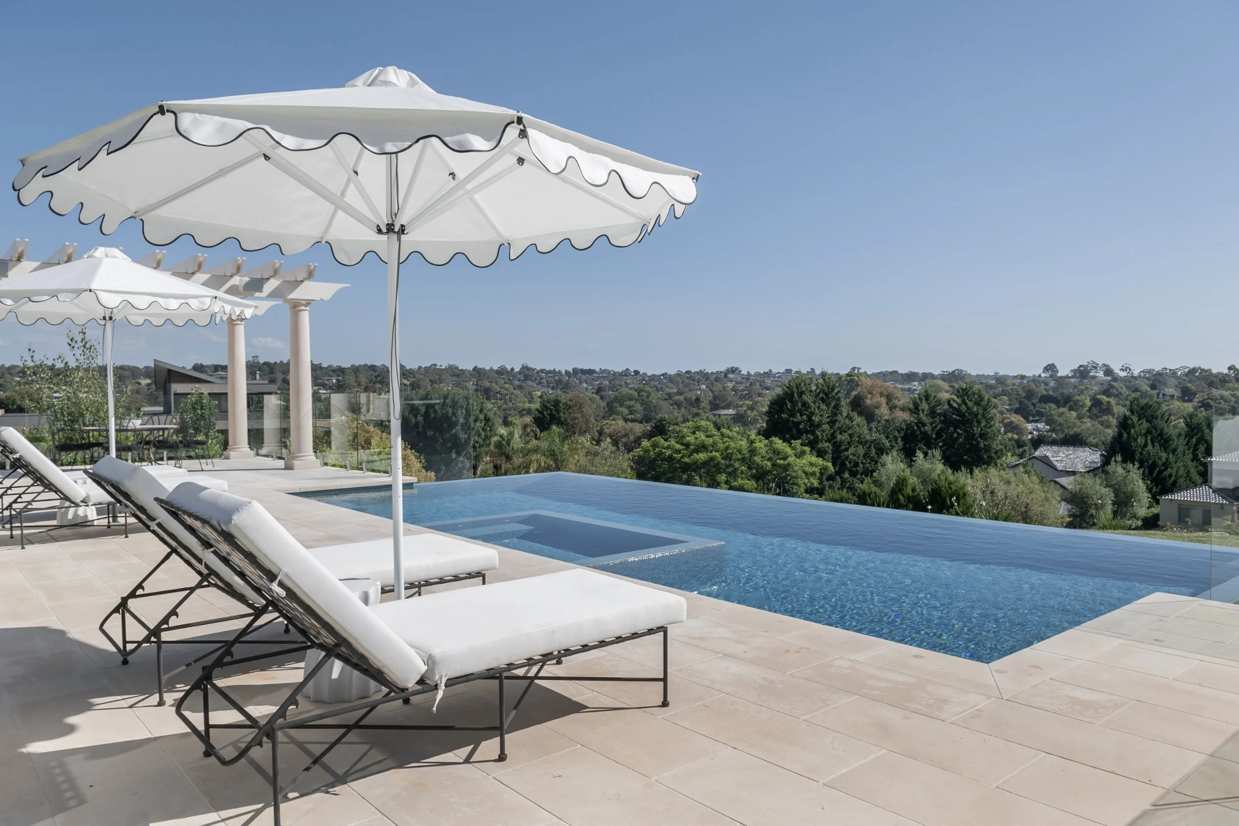Luxury outdoor pool with infinity edge, surrounded by white lounge chairs and umbrellas, overlooking a lush green landscape and distant neighborhood.