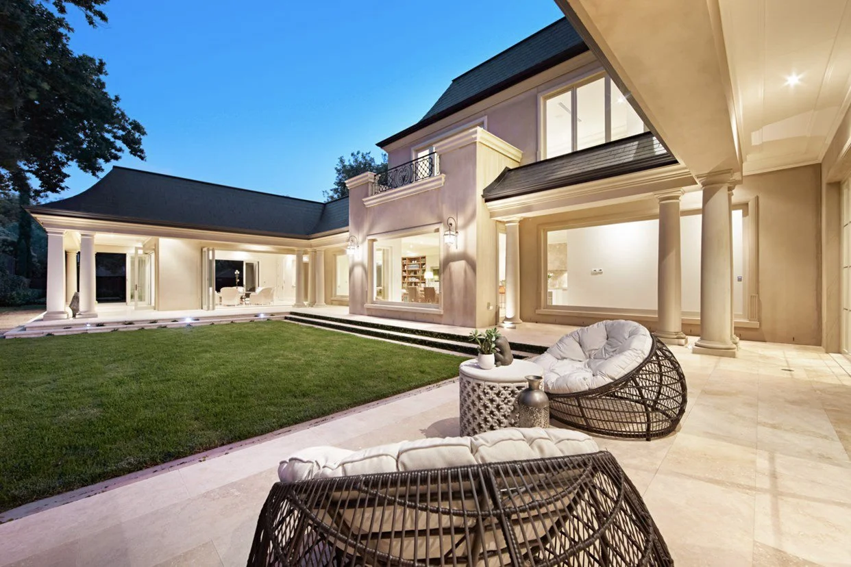 Night view of a large, elegant house with a lit porch, columns, and a well-maintained lawn, furnished with outdoor chairs and a small table.