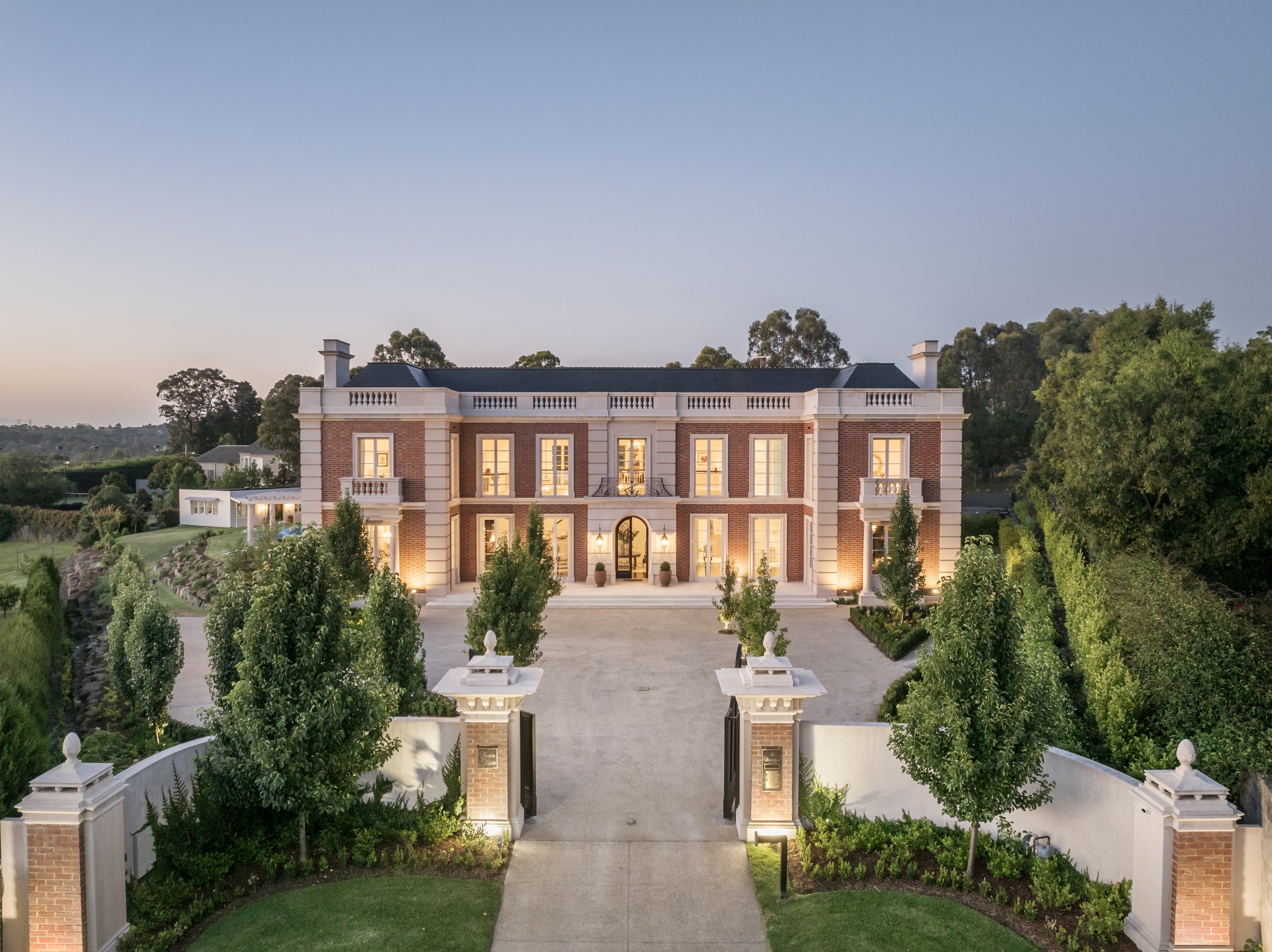 Front view of a large, elegant mansion with brick and white stone exterior, well-lit chateau-style house with a gated entrance, driveway, and lush greenery surrounding the property.