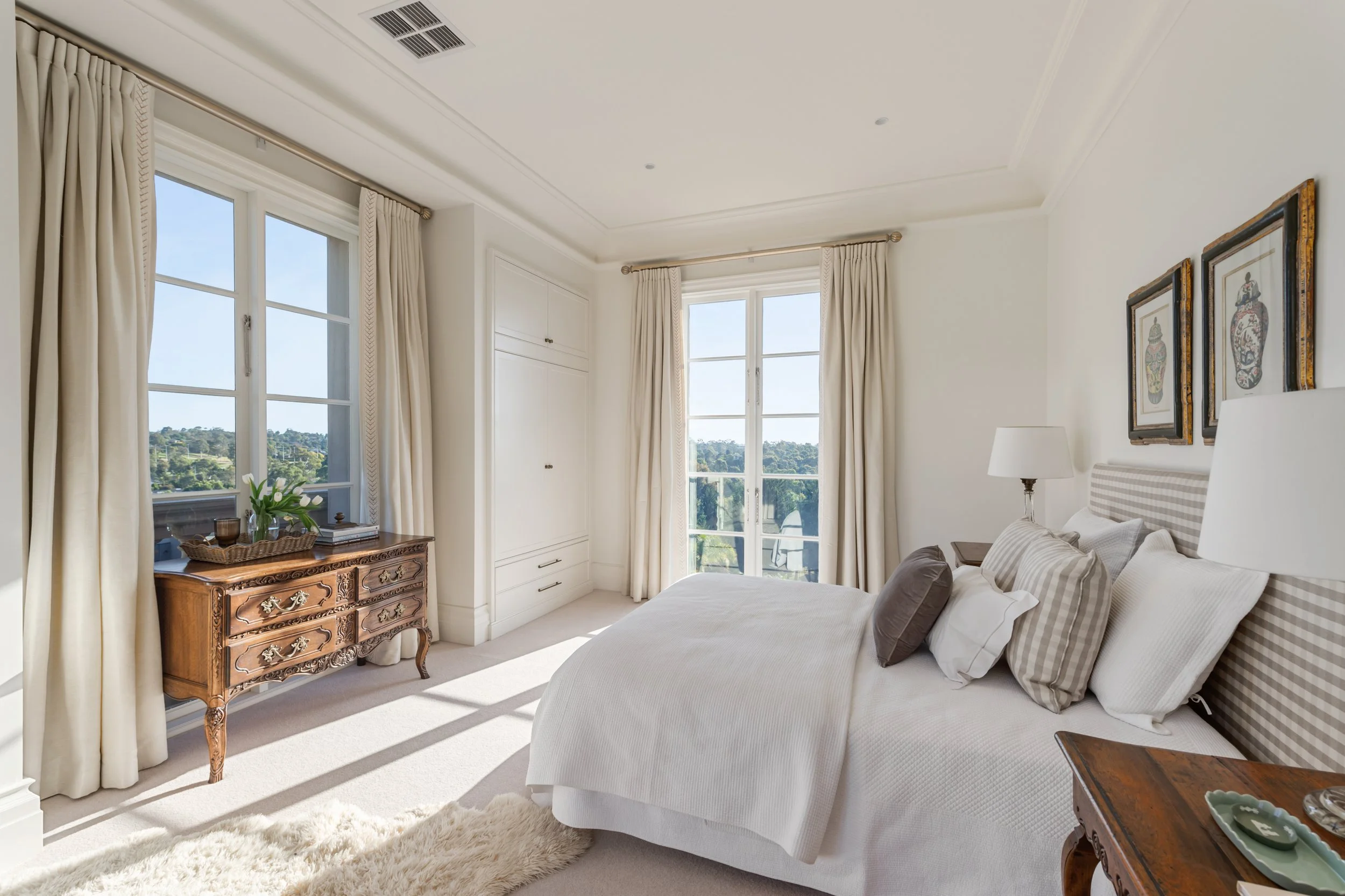 Bright bedroom with white walls, large windows with cream curtains, a bed with white bedding and pillows, wooden side tables, a vintage wooden dresser, framed artwork on the wall, and a cream rug.