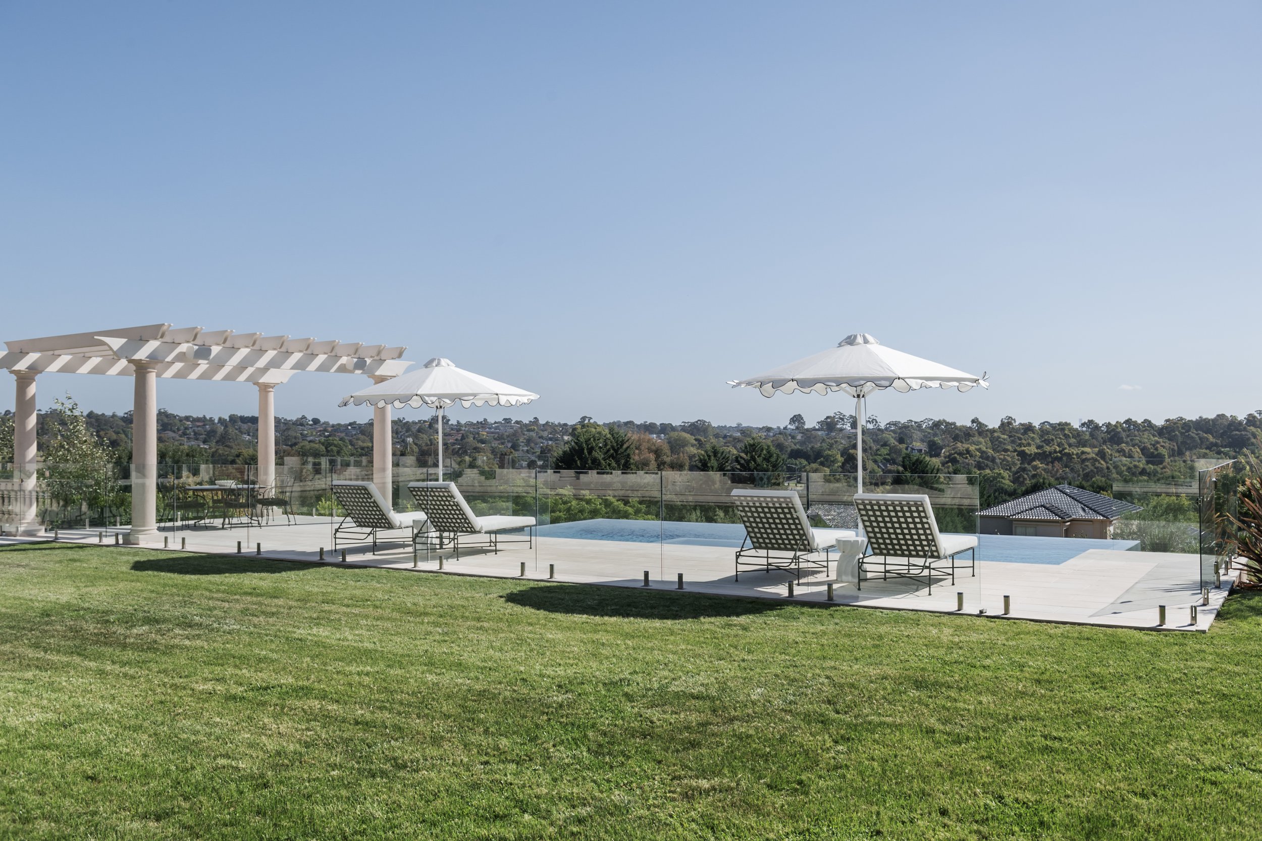 Luxury backyard with a pool, white umbrellas, lounge chairs, and a pergola surrounded by a glass fence, overlooking a lush green landscape under a clear blue sky.
