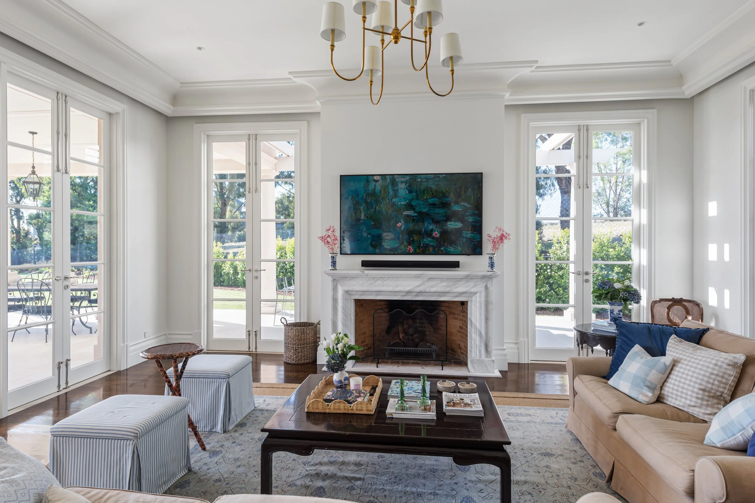 Living room with white walls, large windows, a fireplace with a white mantel, a flat-screen TV above the fireplace, a beige sofa with blue and checkered pillows, a wooden coffee table with decorative items, a striped ottoman, and a chandelier hanging