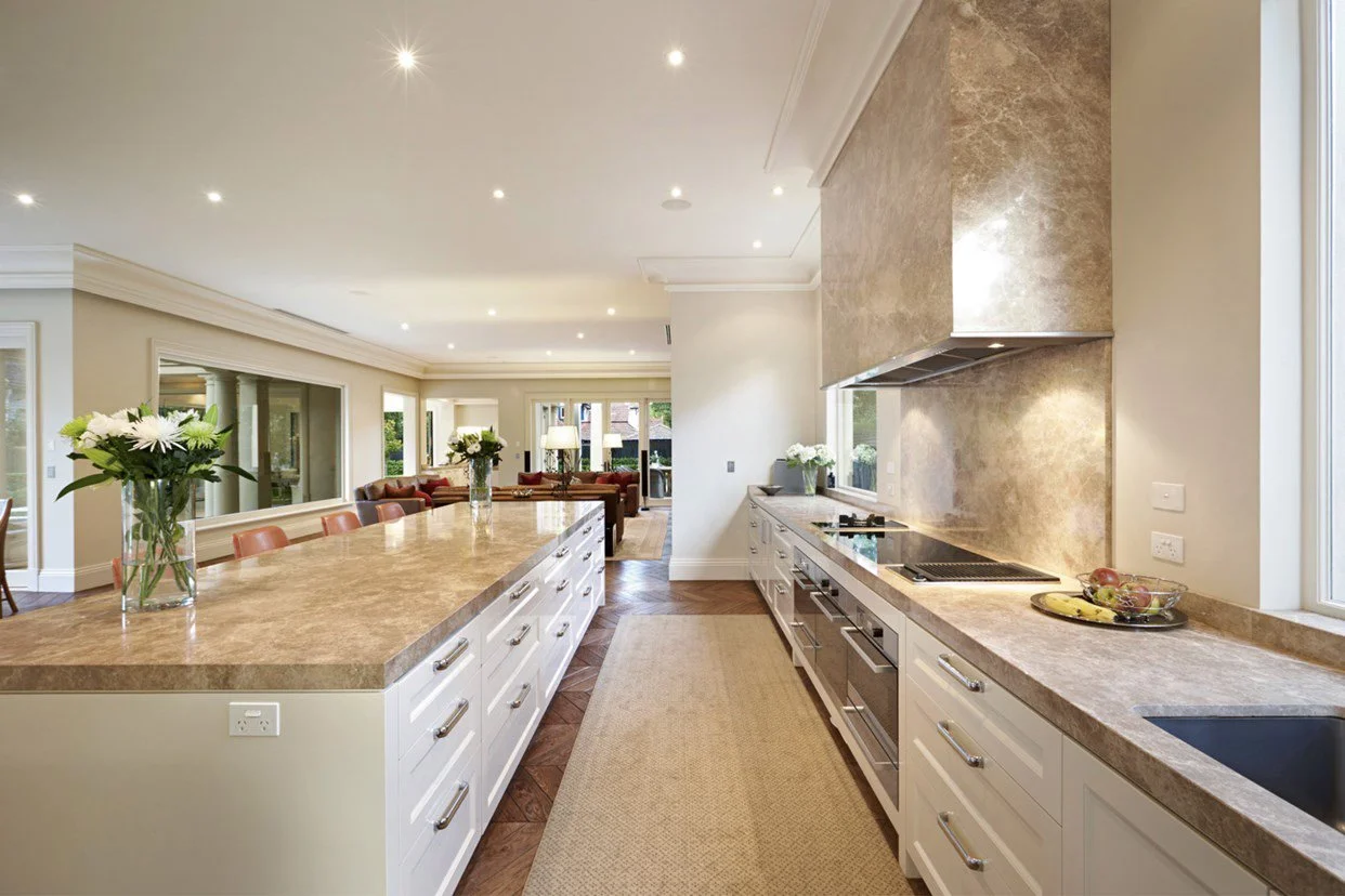 Modern kitchen with a spacious island, white cabinetry, beige countertops, stainless steel appliances, and natural light from large windows, with a view into a living area with seating and decor.