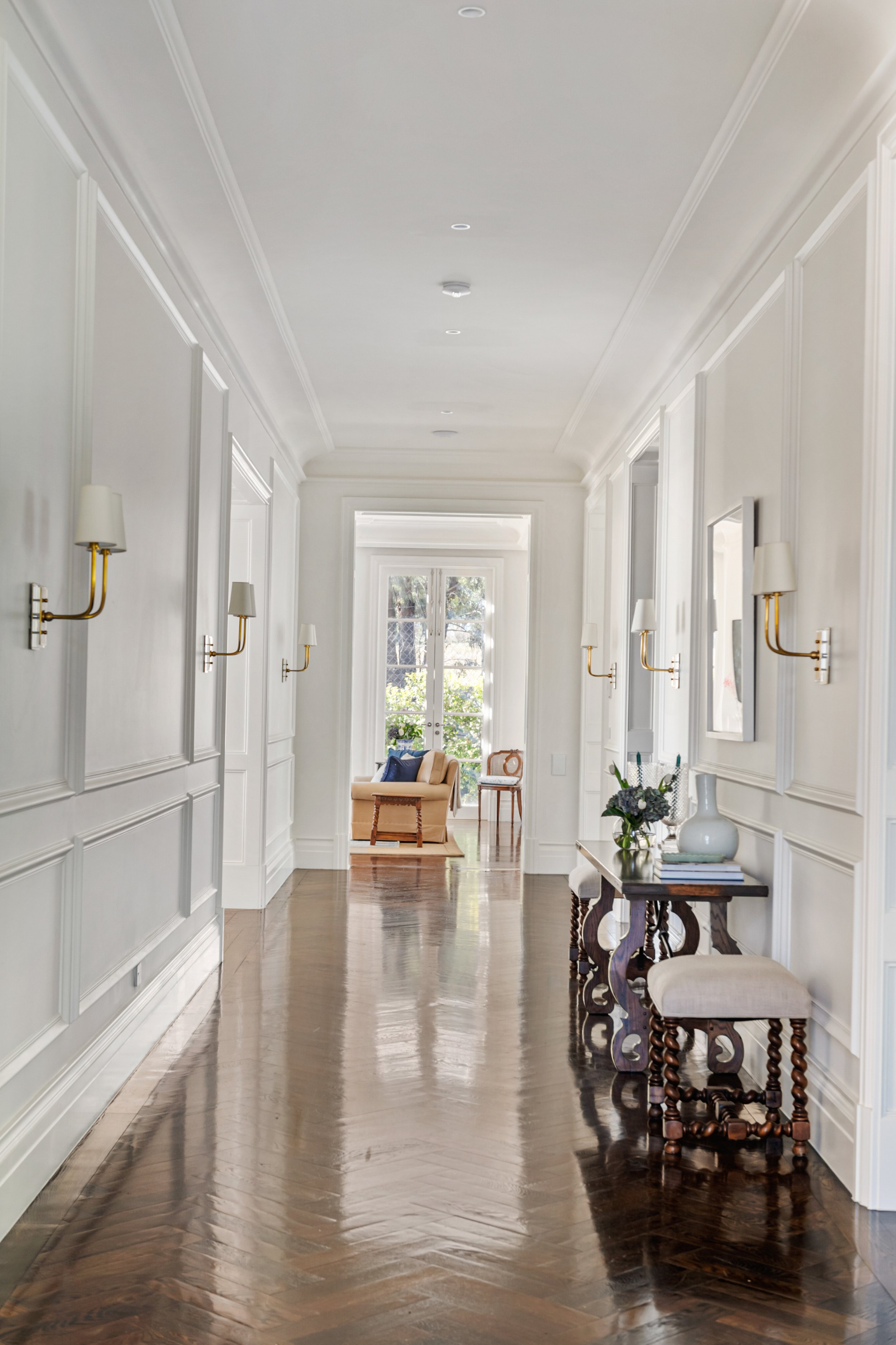 Bright, elegant hallway with white walls, decorative molding, dark wooden flooring, and gold wall sconces. A side table with a vase and books is on the right, leading to a cozy sitting area with a beige armchair and large window in the background.
