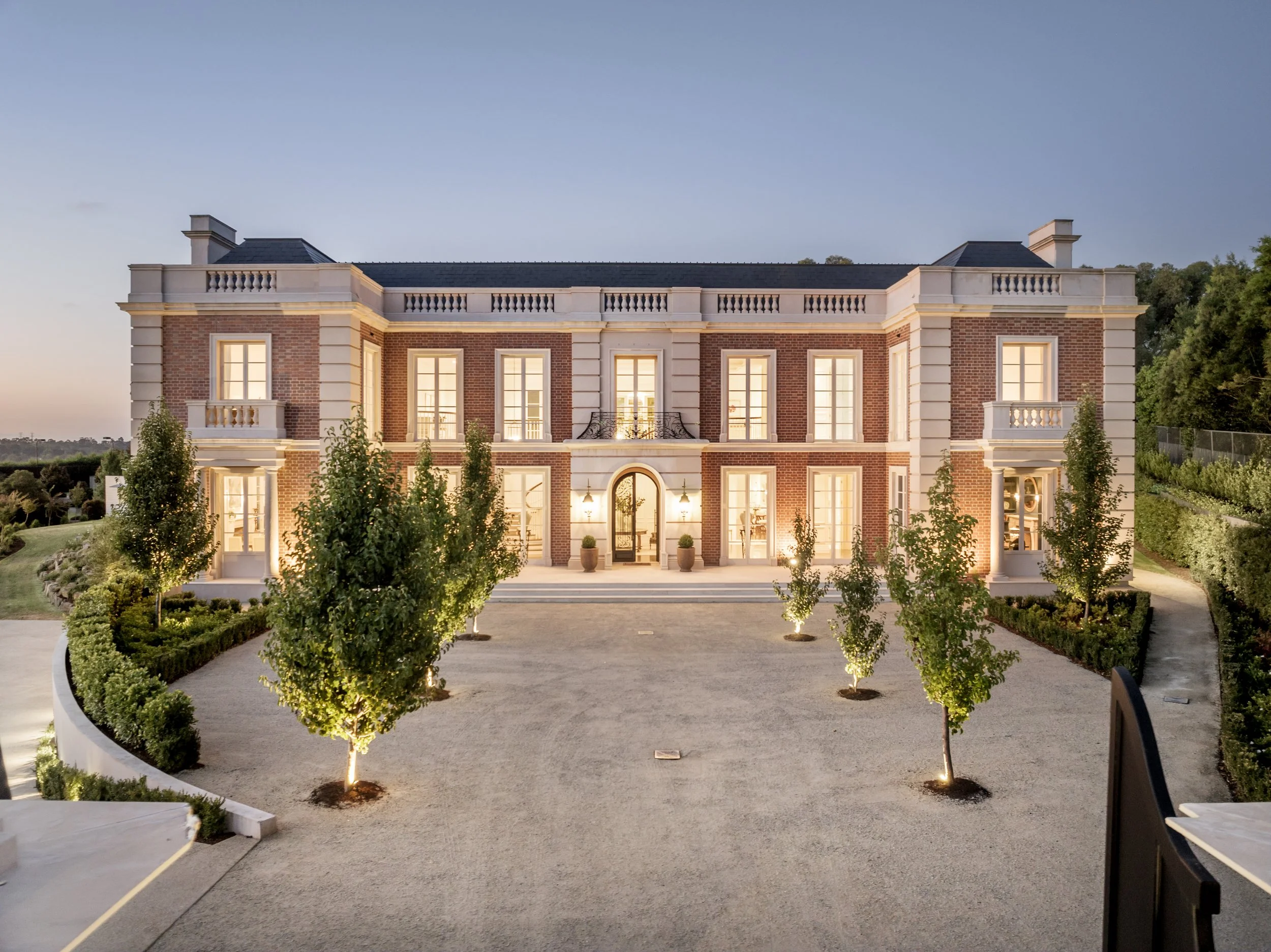 Elegant three-story mansion with brick facade, white accents, and large illuminated windows, surrounded by landscaped garden with trees and low hedges.