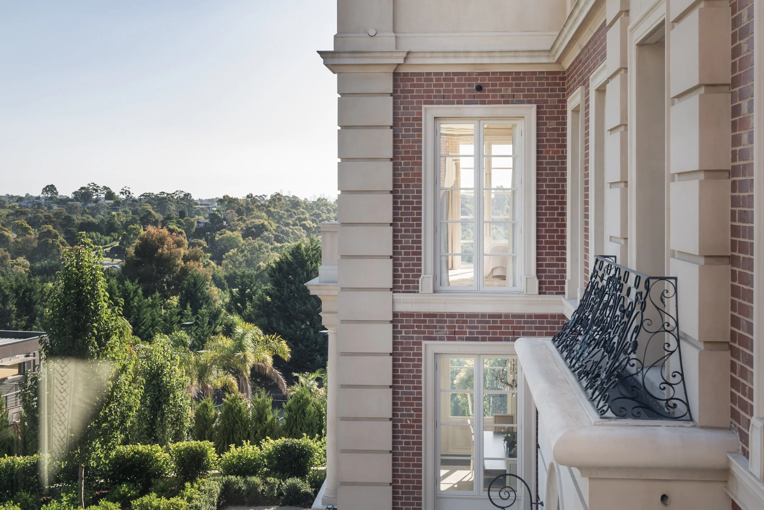 Corner of a brick and cream-colored building with multiple windows, ornate iron balcony, and lush green trees and hills in the background.