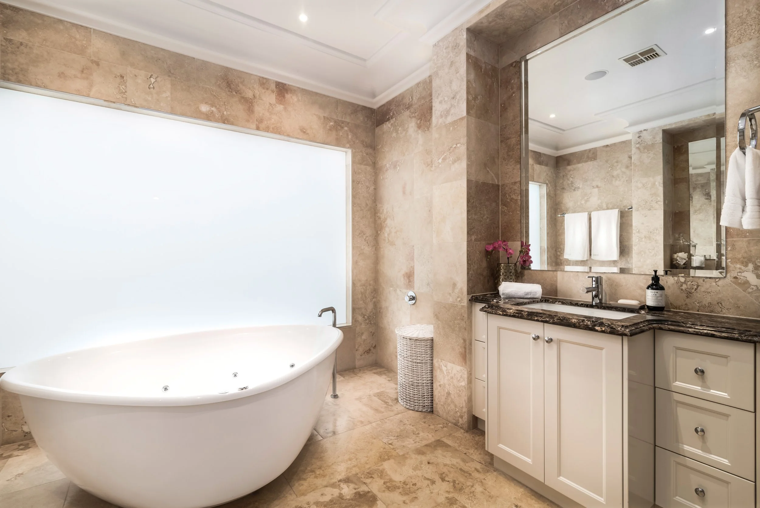 A luxurious bathroom with a large white bathtub, beige marble walls and floors, a vanity with a dark granite countertop, a large mirror, towels, pink flowers, and a soap dispenser.