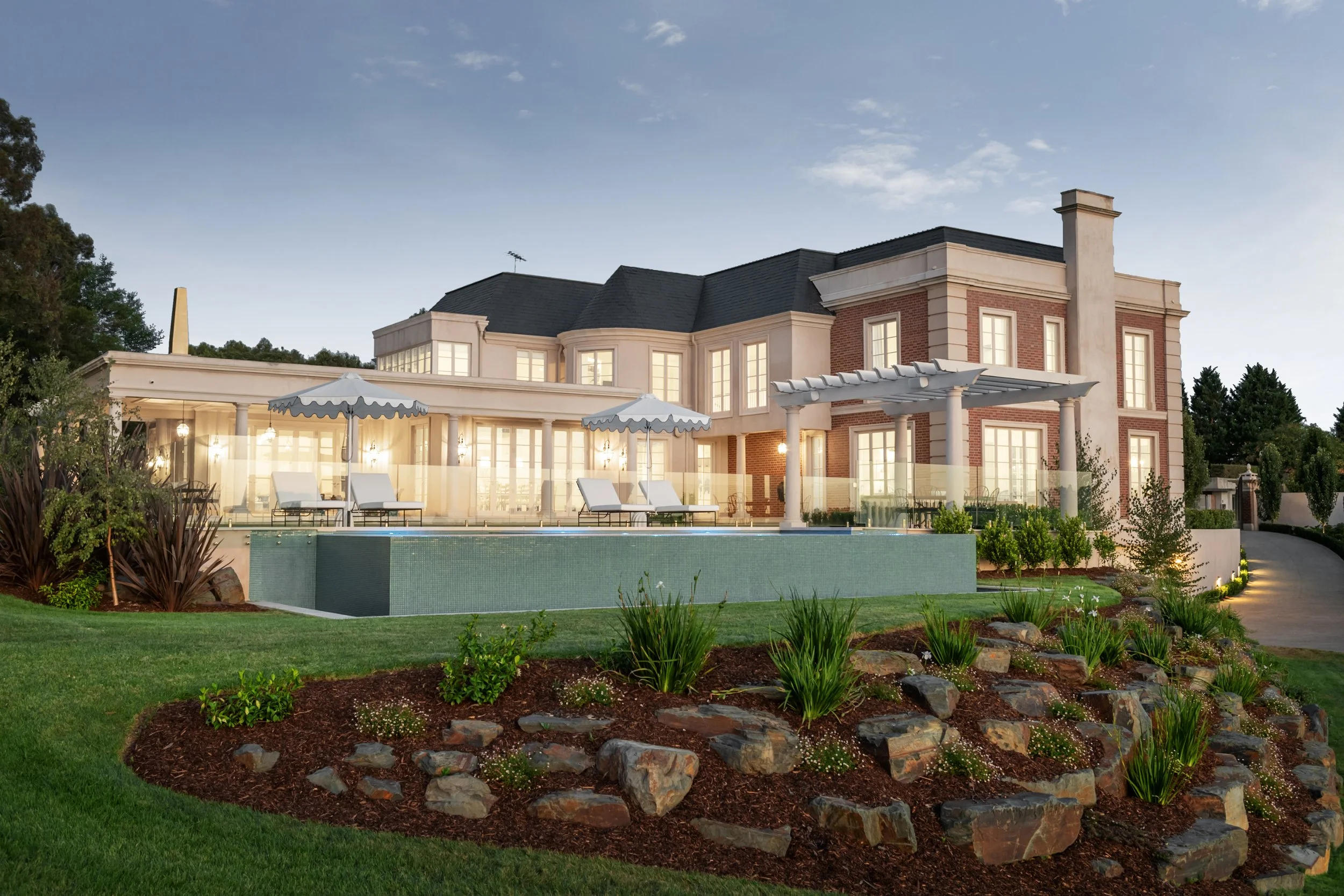 Luxury house with a swimming pool, outdoor lounge chairs with umbrellas, manicured lawn, landscaped garden with rocks and plants, and a curved driveway at dusk.