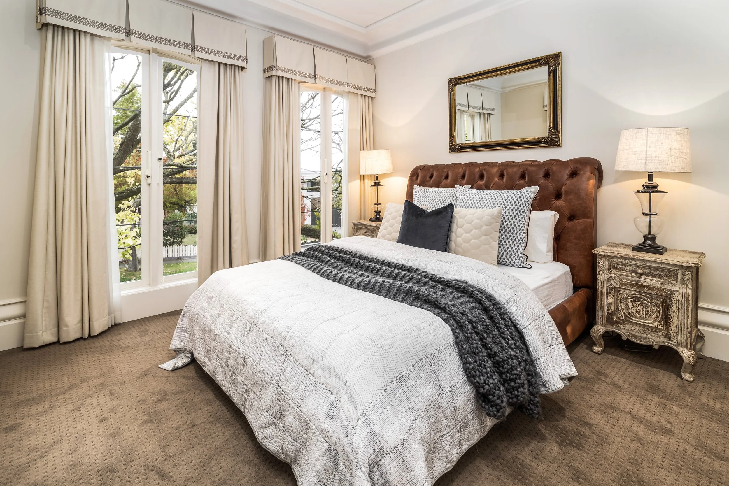 A cozy bedroom with a large bed that has a leather headboard, multiple decorative pillows, and a textured gray blanket. There are two vintage-style nightstands with lamps on each side of the bed. Large windows with cream-colored curtains let in natur