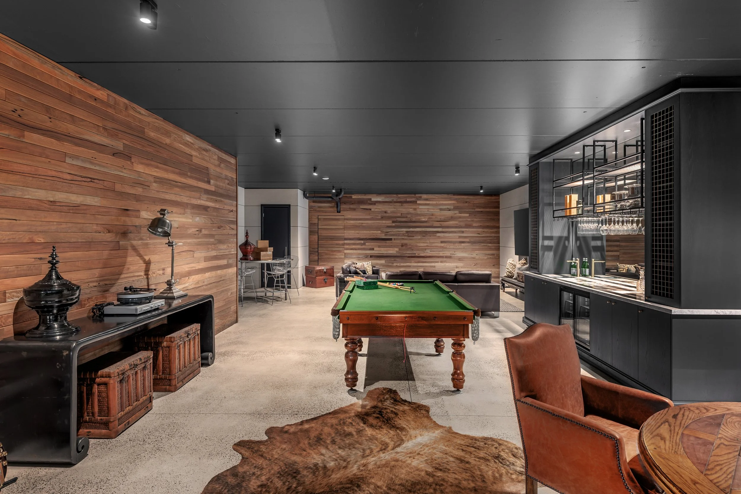 A recreational room with a pool table in the center, a leather sofa, a bar area with glasses, a cowhide rug, and wooden wall paneling.