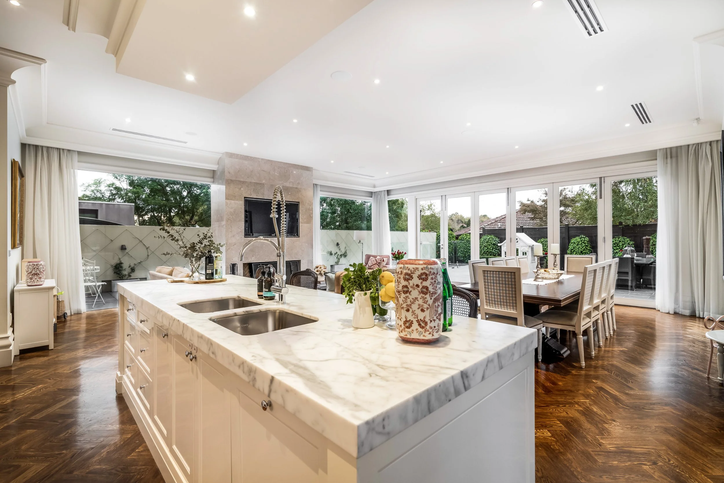 Modern open-concept kitchen and dining area with white marble island, hardwood floors, large windows, and a view of a backyard with trees and outdoor seating
