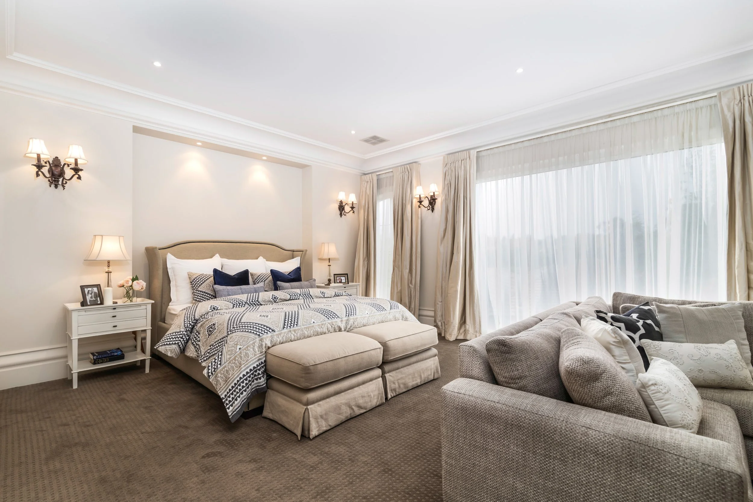 A spacious bedroom with a bed, side tables, lamps, and a beige sofa with throw pillows, large windows with sheer curtains, and elegant wall sconces.