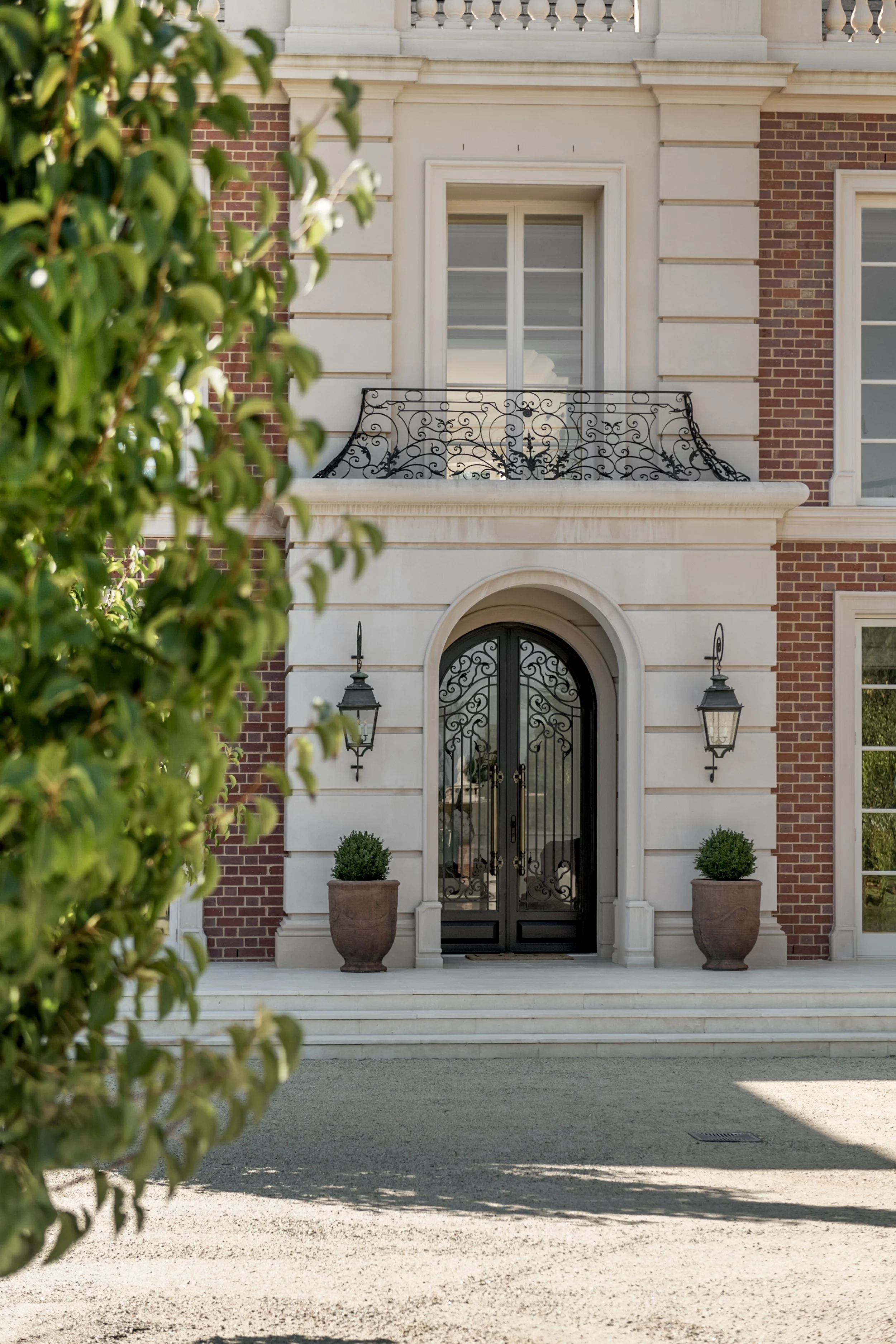 Front view of a grand house with a black wrought iron door flanked by two large potted plants and two wall-mounted lanterns, with a second-story balcony and windows behind, partially obscured by greenery.
