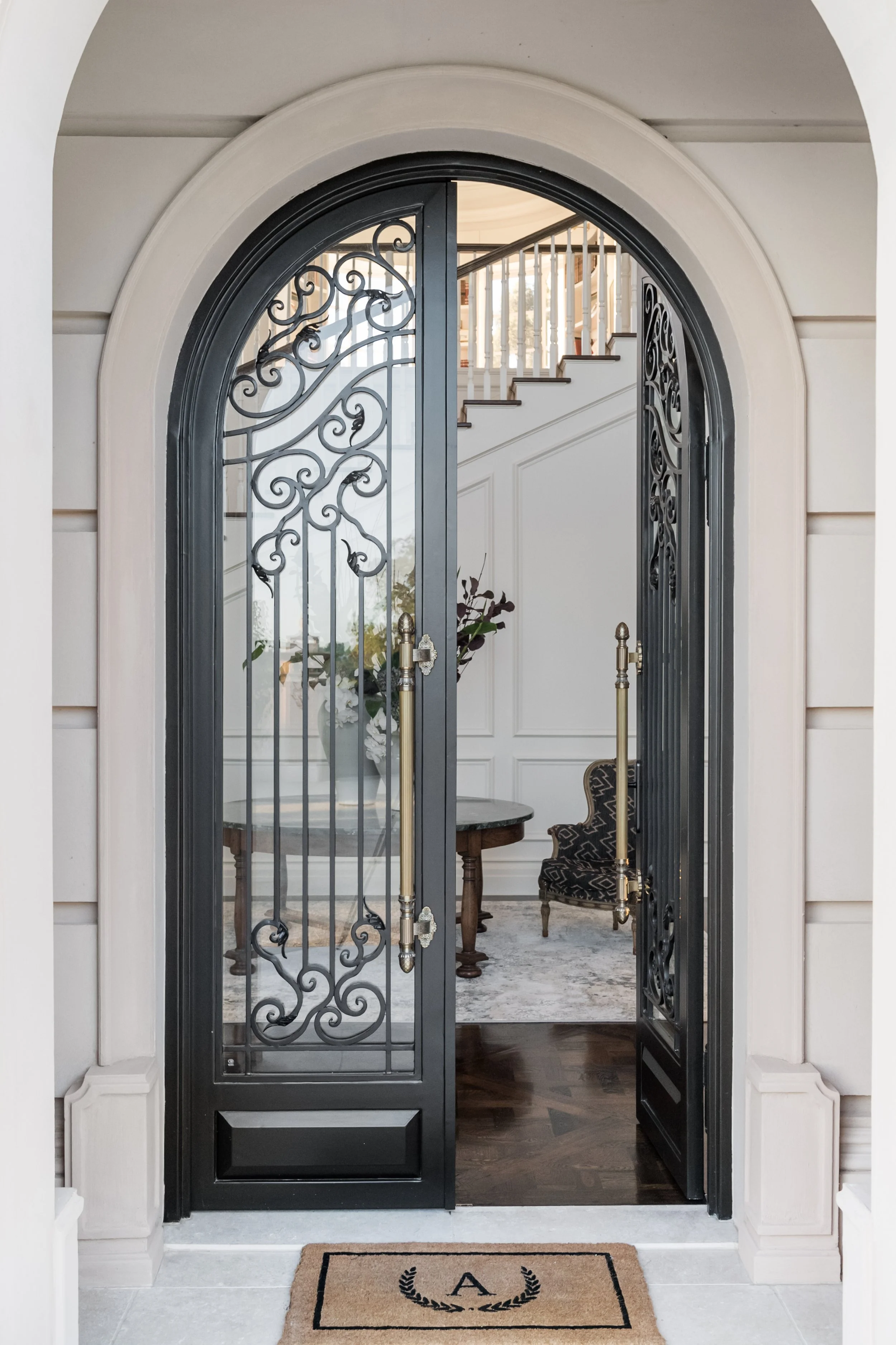 Black wrought iron security door with decorative scrollwork, opening into a foyer with staircase, side chair, and table with flowers.